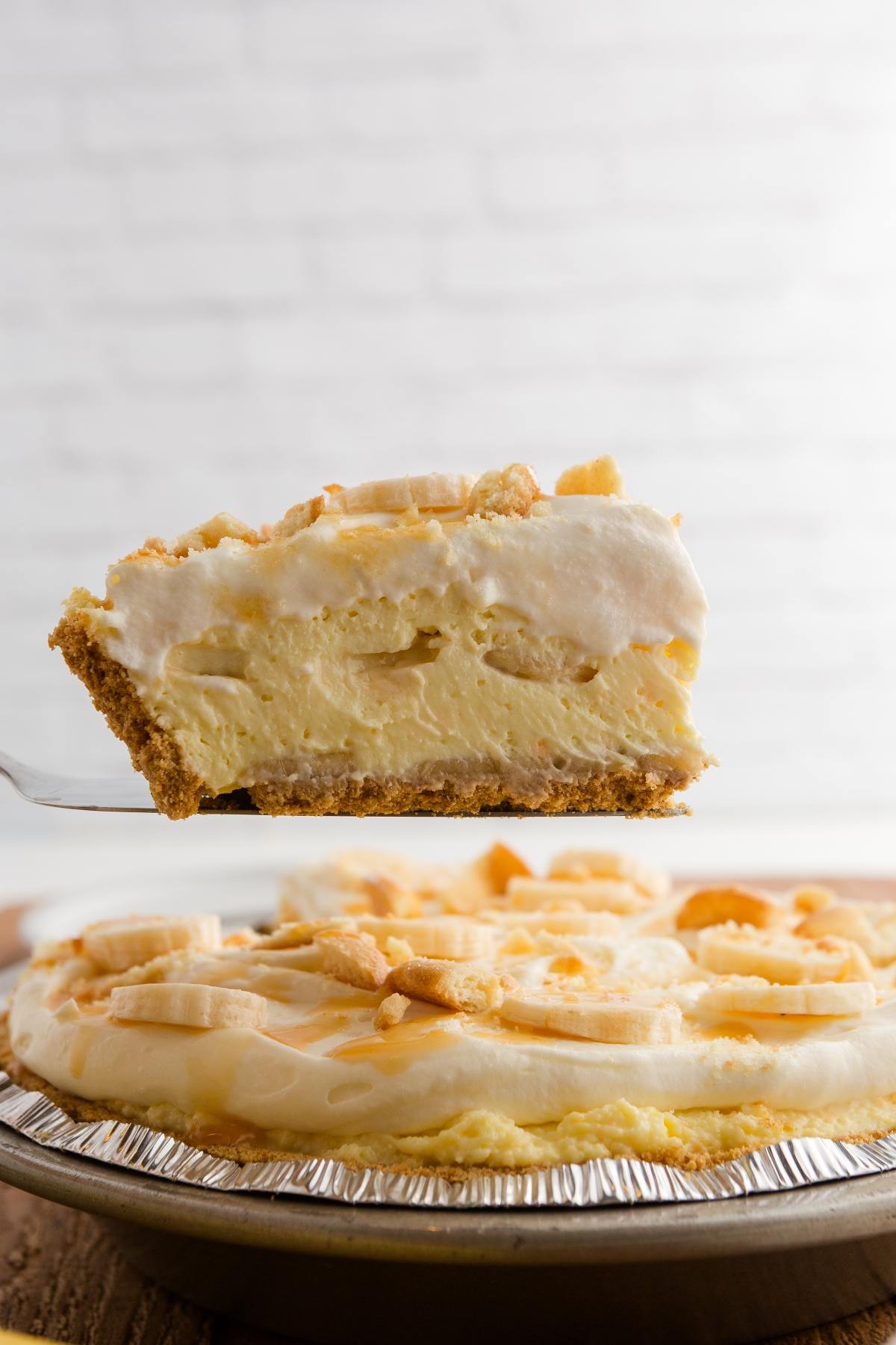 A slice of creamy banana pudding pie with a graham cracker crust is lifted above the whole pie.