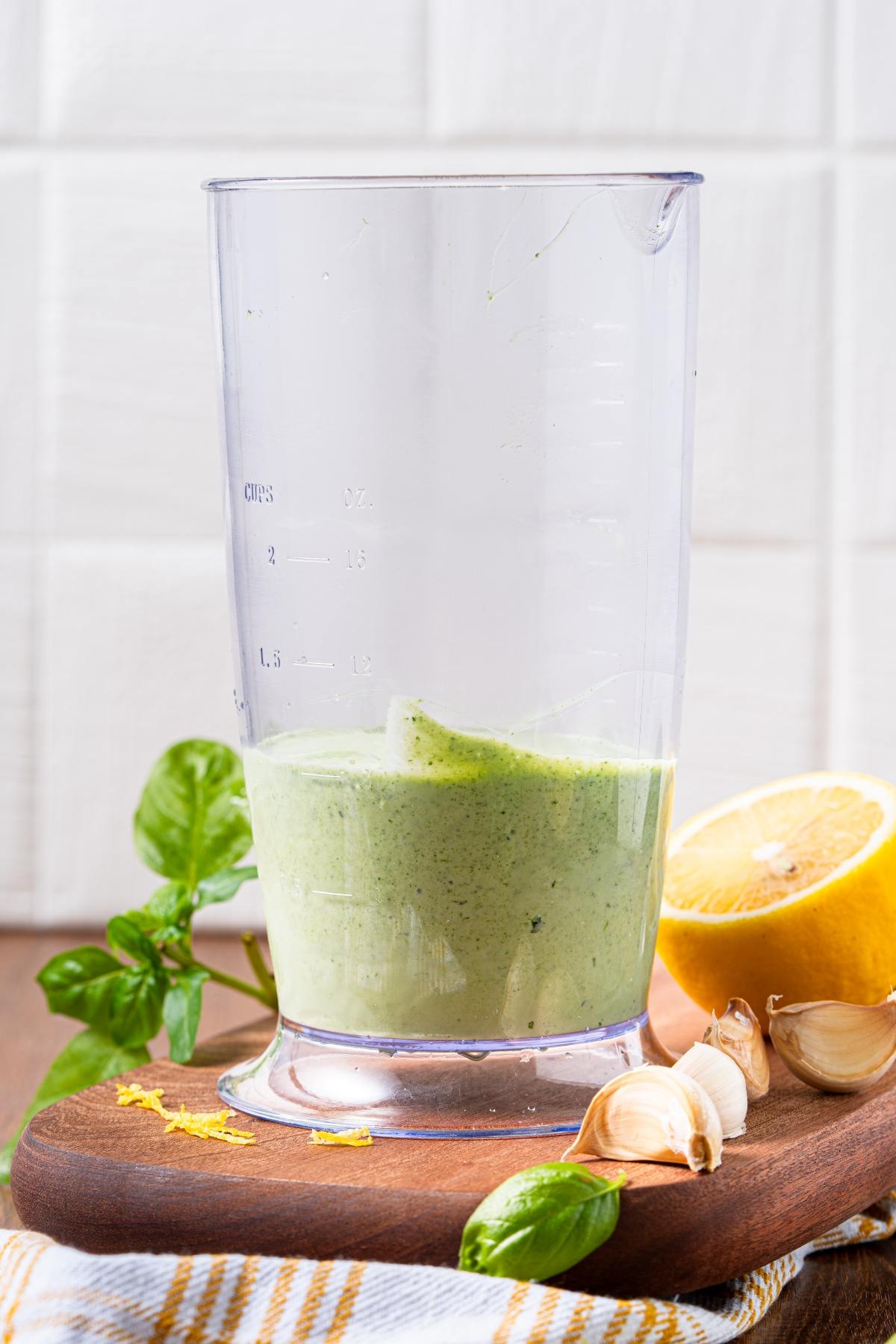 A glass measuring cup with green sauce, surrounded by basil, garlic, and a halved lemon on a wooden board.