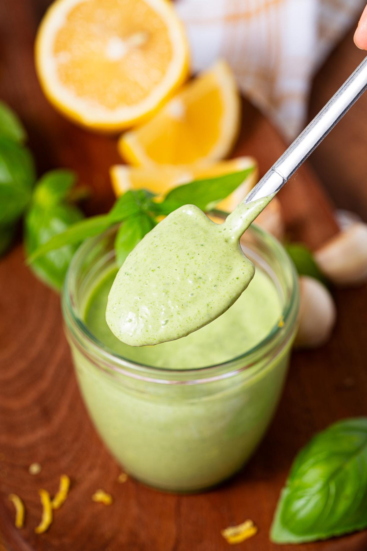 A spoon holding creamy green sauce above a glass jar, with lemon and basil in the background.