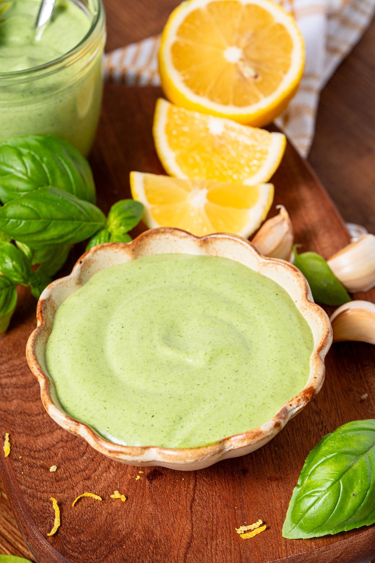 A bowl of creamy green sauce with lemon wedges, basil leaves, and garlic on a wooden board.
