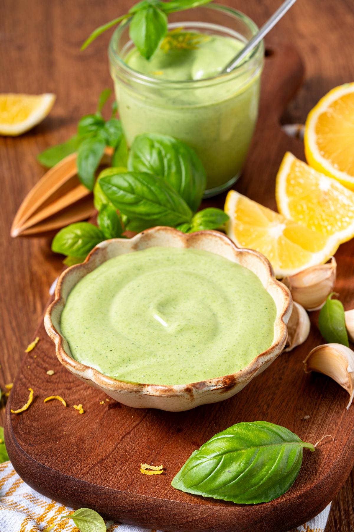 Creamy green sauce in a bowl, surrounded by basil leaves, lemon wedges, garlic, and a jar of more sauce.