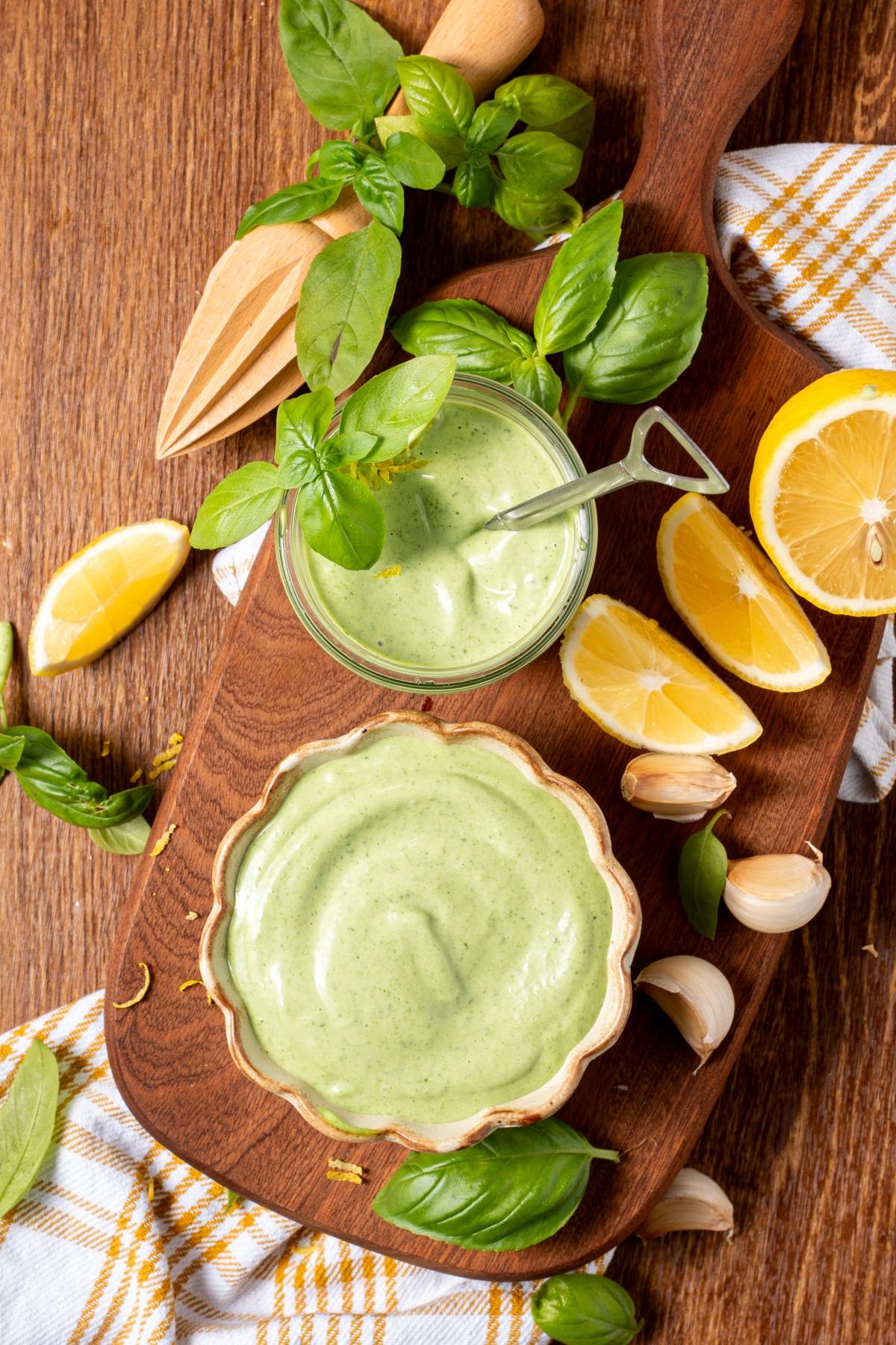 Two bowls of green sauce on a wooden board with basil, lemon wedges, garlic, and a kitchen towel.
