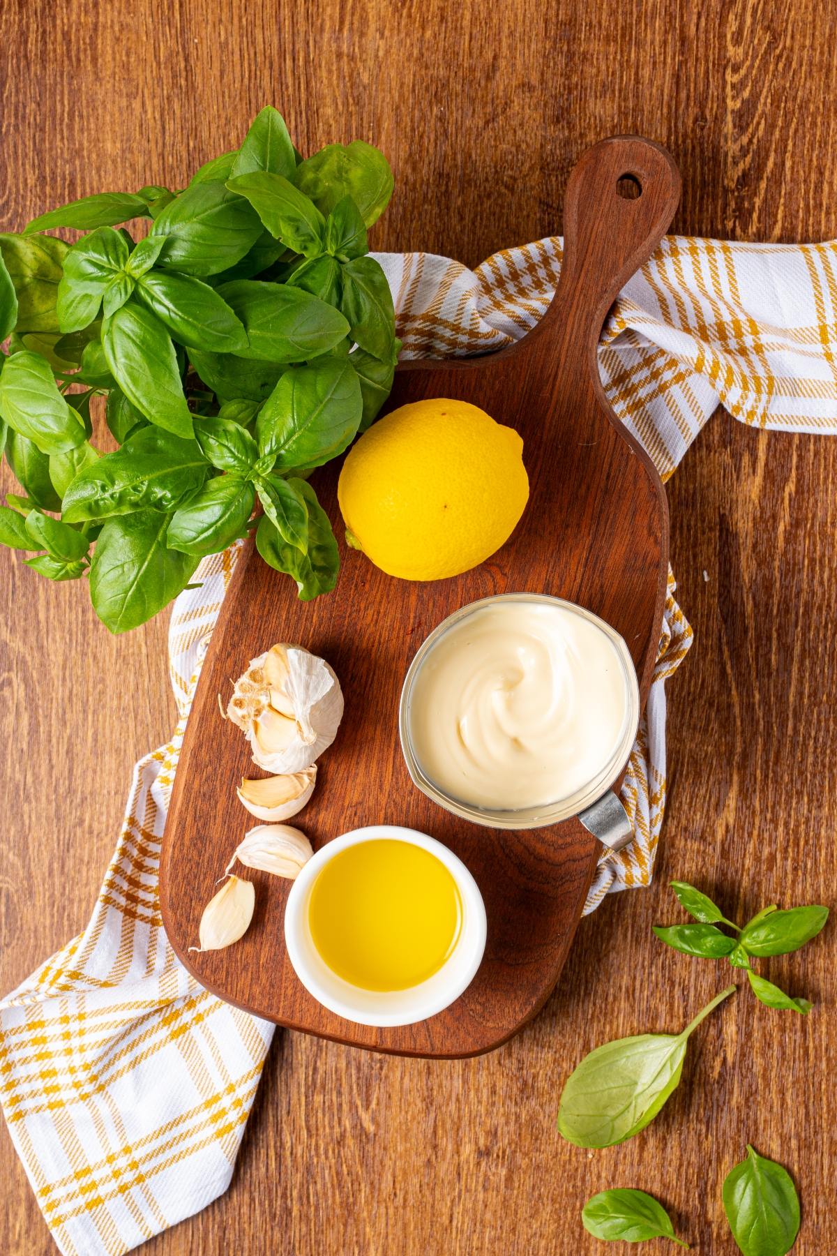 A wooden board with basil, a lemon, garlic, mayonnaise, and olive oil on a checked cloth.