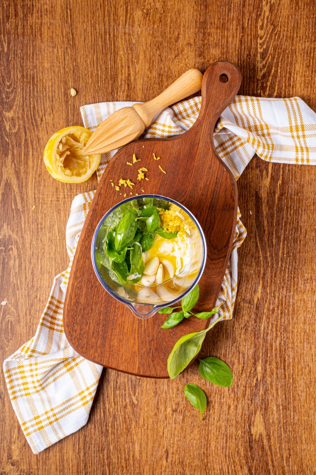 A bowl with basil leaves, lemon zest, and mayonnaise on a wooden board with a citrus reamer and cloth.