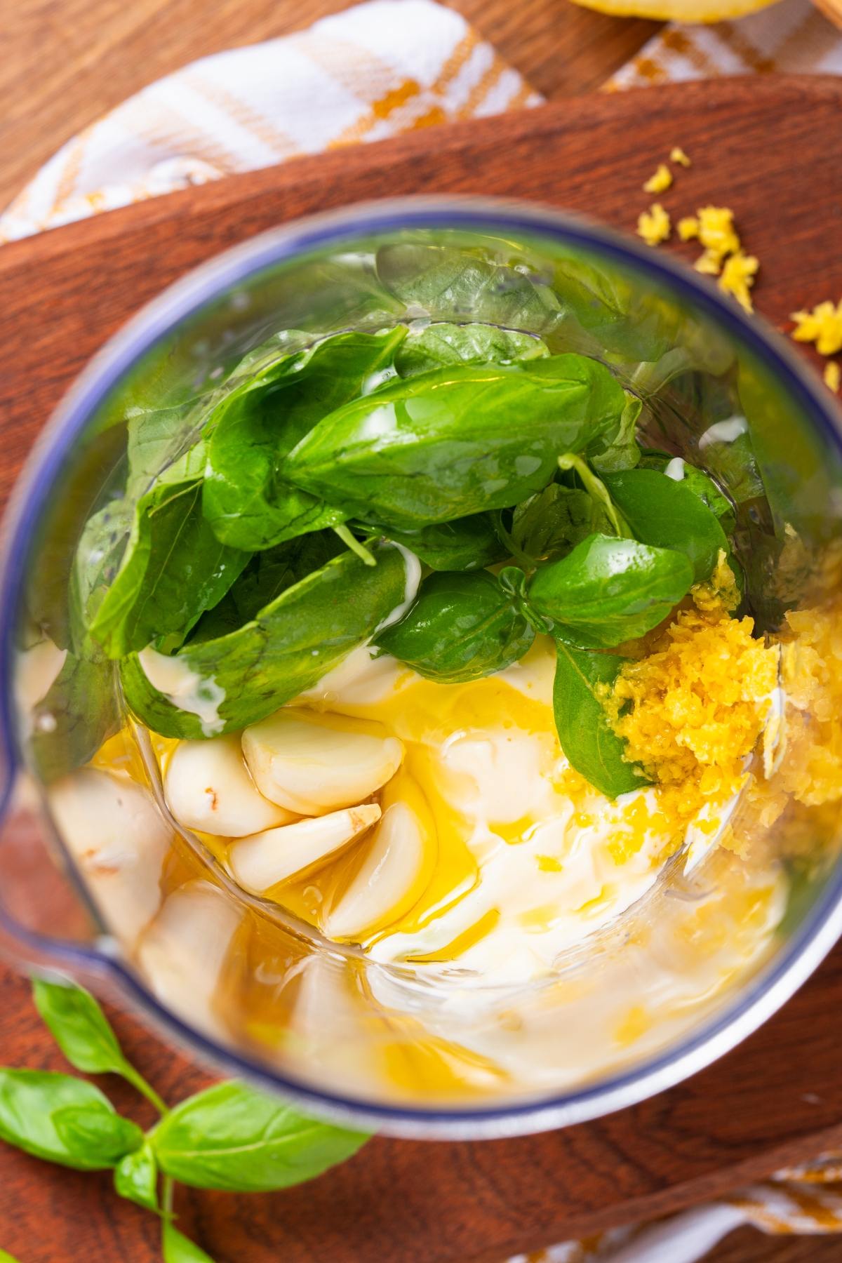 Fresh basil, garlic cloves, lemon zest, and oil in a blender, ready to be mixed for a sauce or dressing.