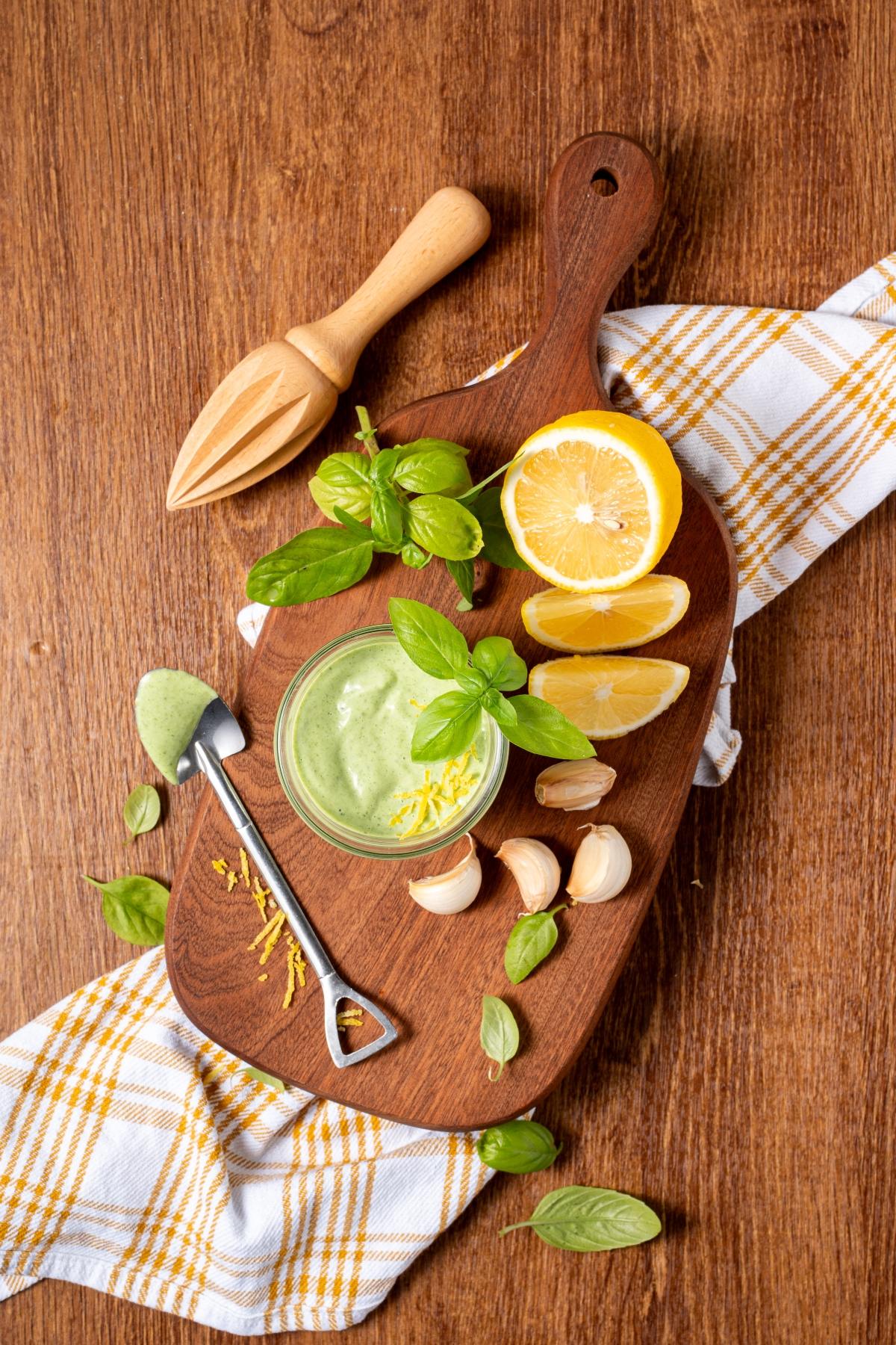 A jar of green sauce, garlic, lemon halves, basil, and a citrus juicer on a wooden board with a towel.