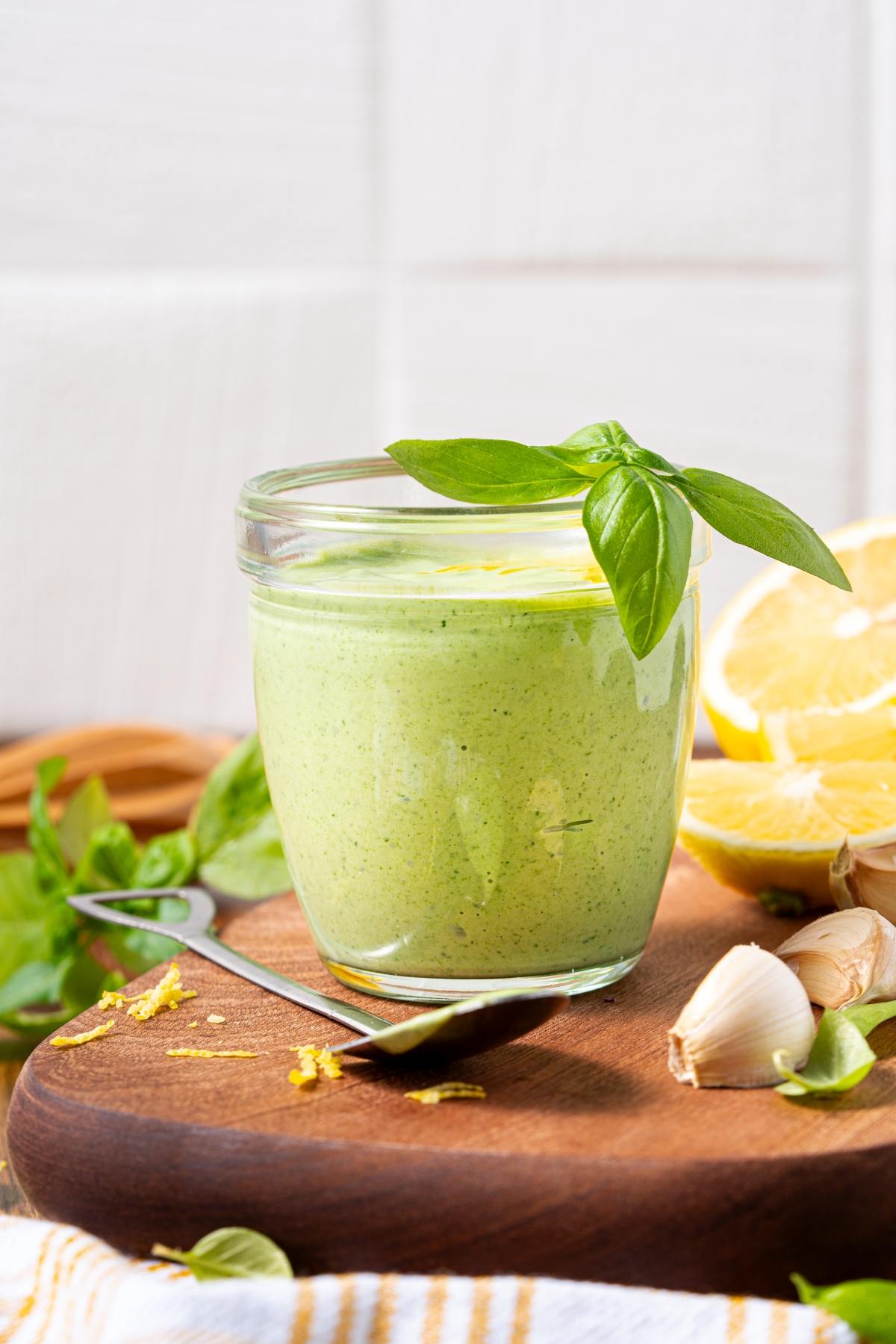 A jar of green sauce garnished with basil, beside lemon wedges, garlic, and a spoon on a wooden board.