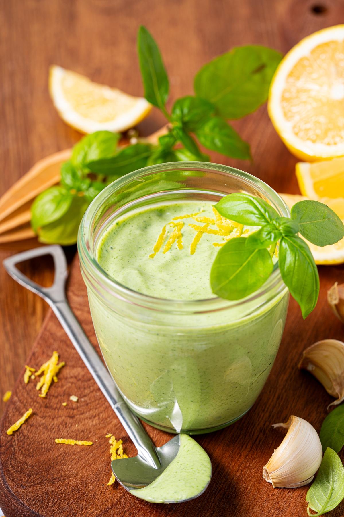 A glass jar of green sauce garnished with lemon zest and basil, surrounded by lemon, basil, and garlic.