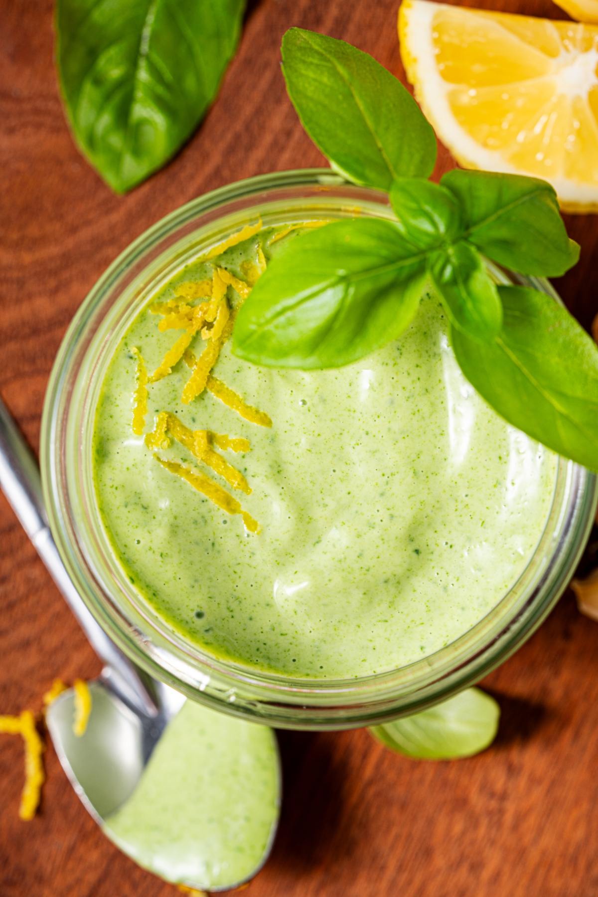 A jar of creamy green sauce garnished with basil leaves and lemon zest, with a spoon and lemon wedge nearby.