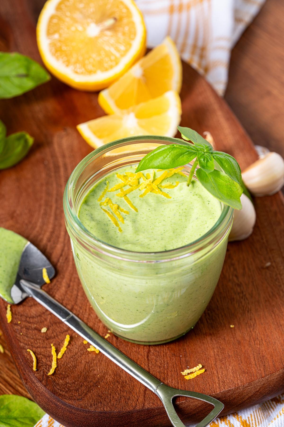 A jar of green sauce topped with lemon zest and basil, with lemon slices and garlic on a wooden board.