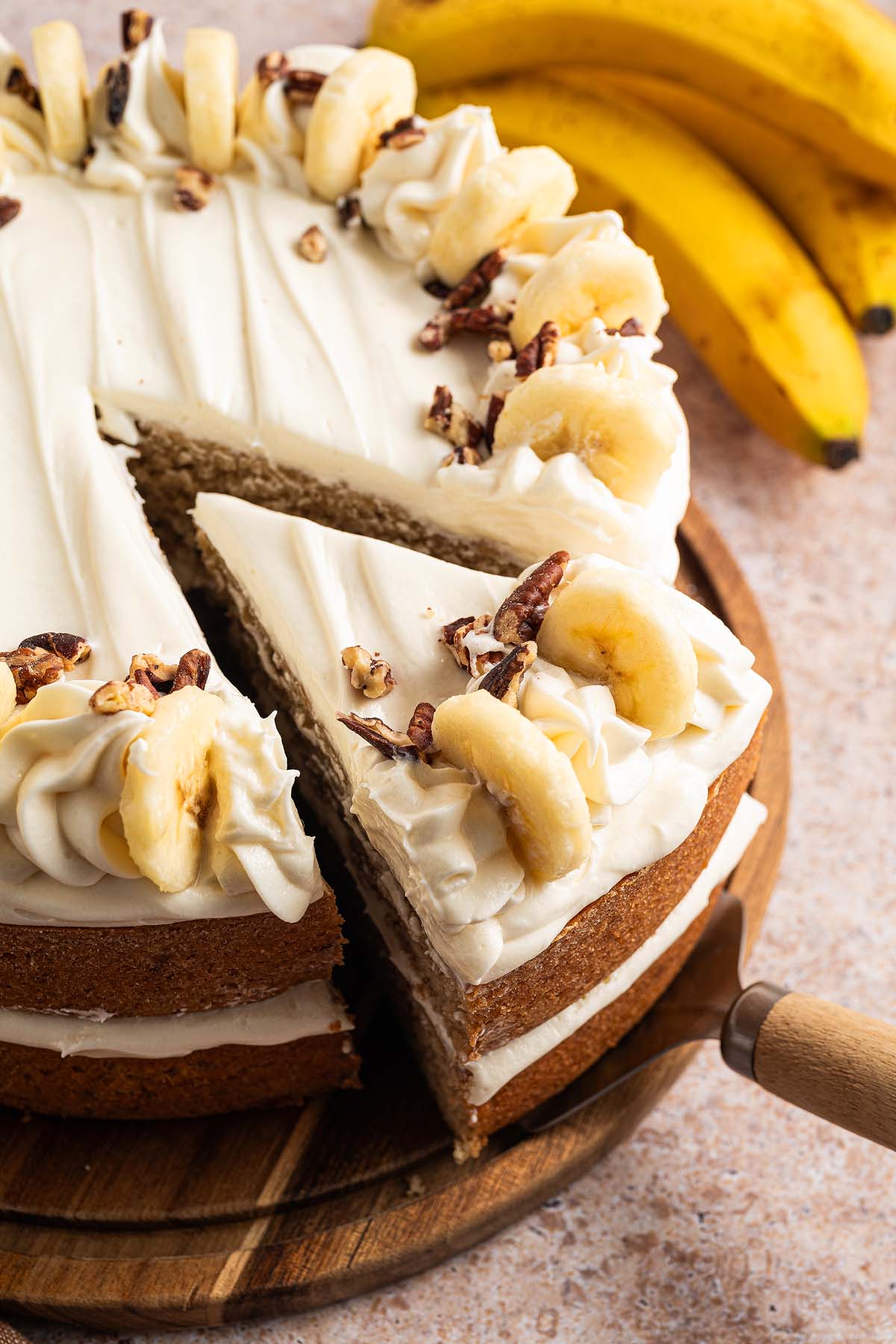 Banana Cake with white frosting, banana slices, and nuts, with a slice being lifted; ripe bananas in the background.