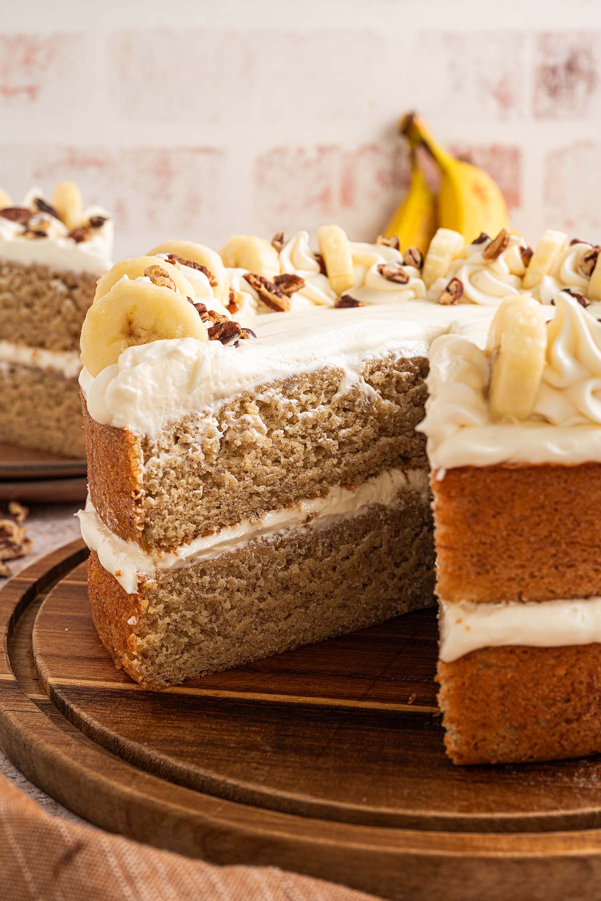 A delectable Banana Cake with white frosting and banana slices sits on a wooden plate, with fresh bananas in the background.