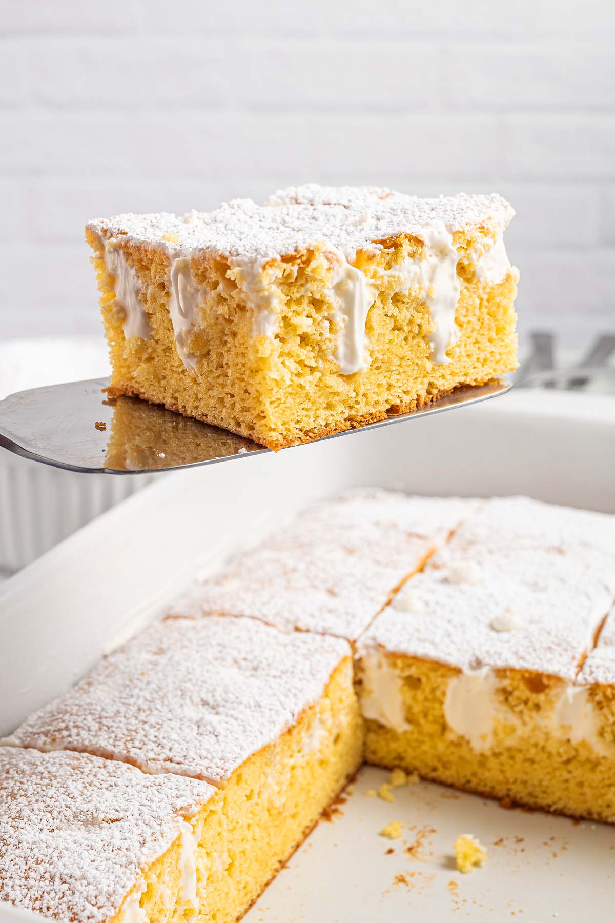 A slice of Twinkie Poke Cake with white icing and powdered sugar is being lifted from a baking dish.