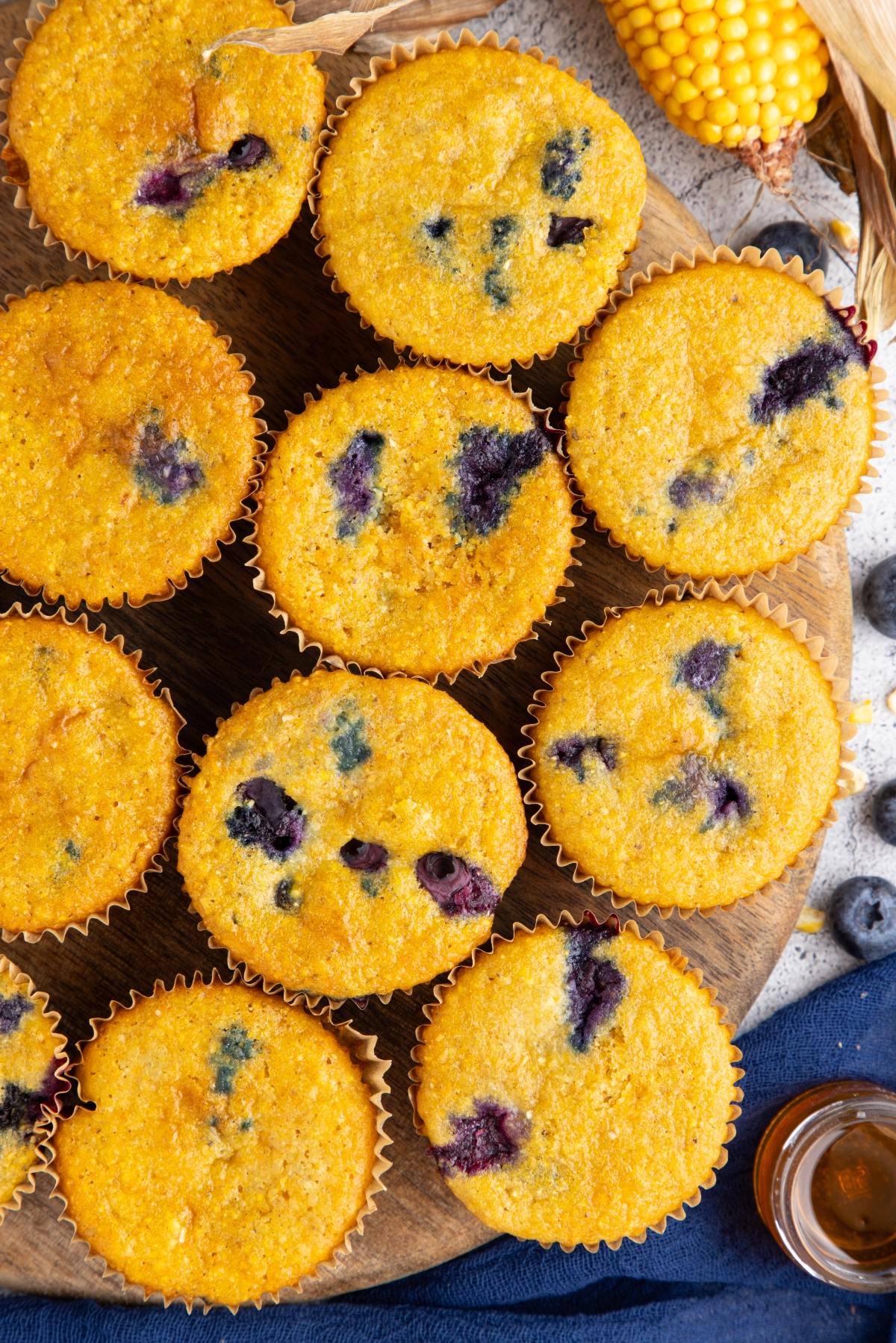 Top view of blueberry cornbread muffins on a wooden board, surrounded by corn and fresh blueberries.