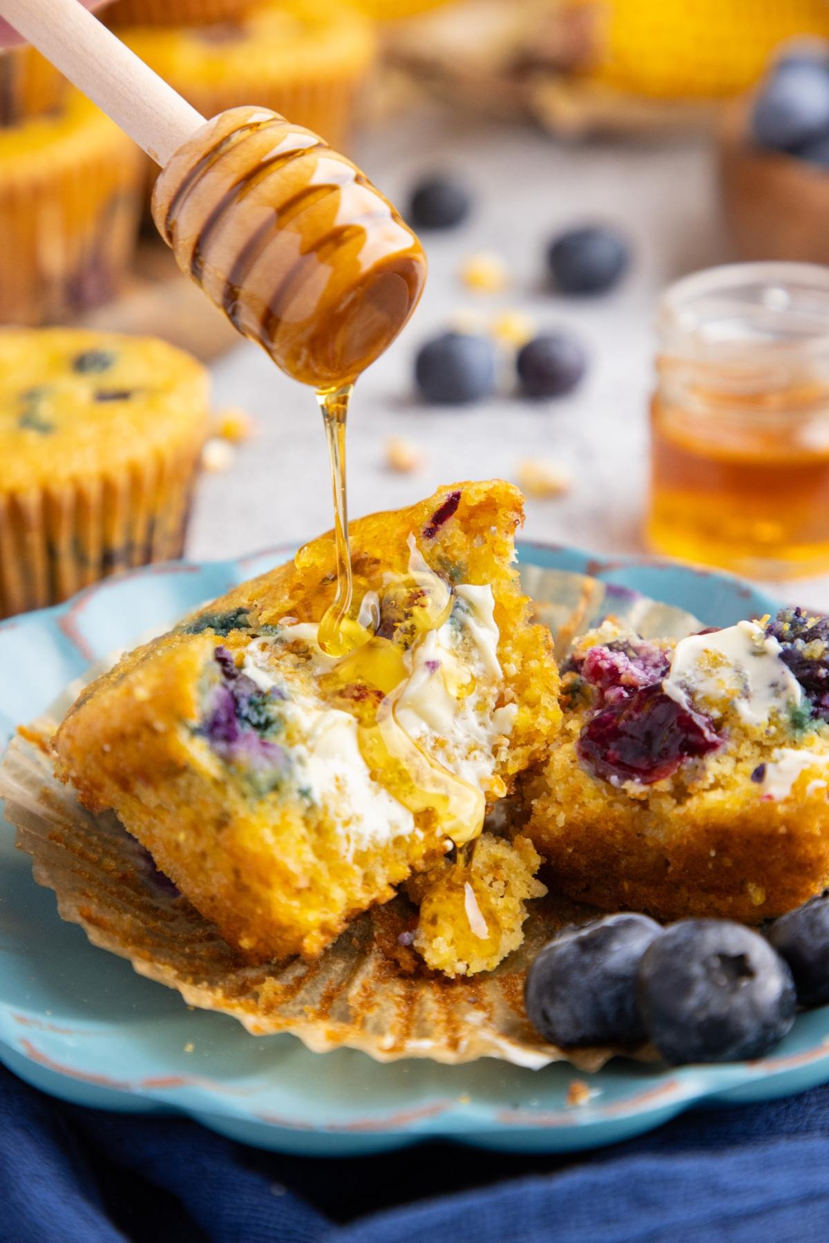 Honey drizzled on a halved blueberry muffin with butter, on a blue plate with fresh blueberries.