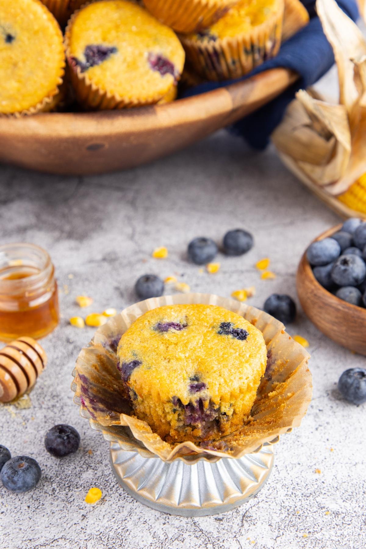A blueberry muffin with a bite taken out, surrounded by blueberries and honey on a textured surface.
