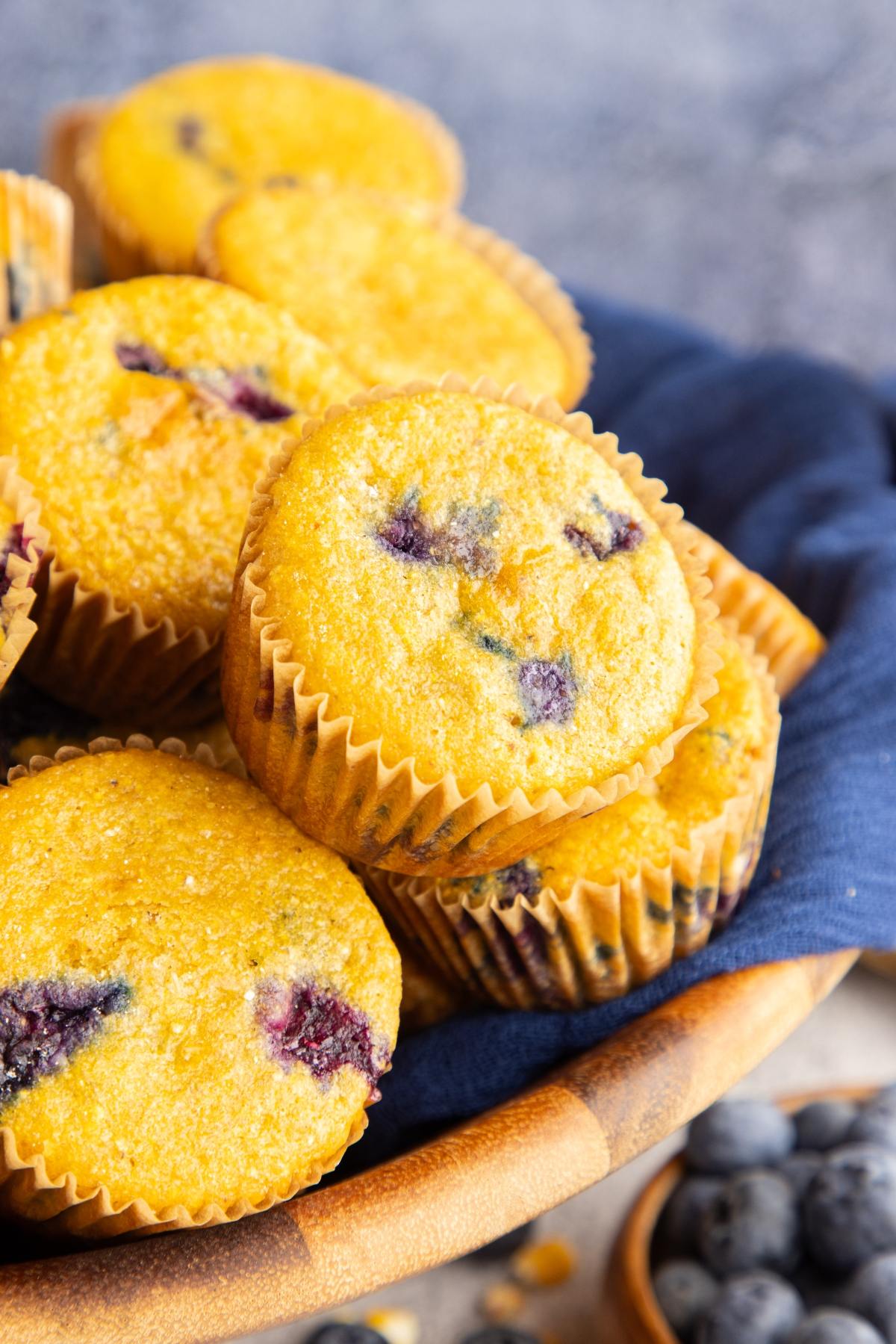 A basket filled with blueberry muffins, some stacked, with a blue cloth underneath.
