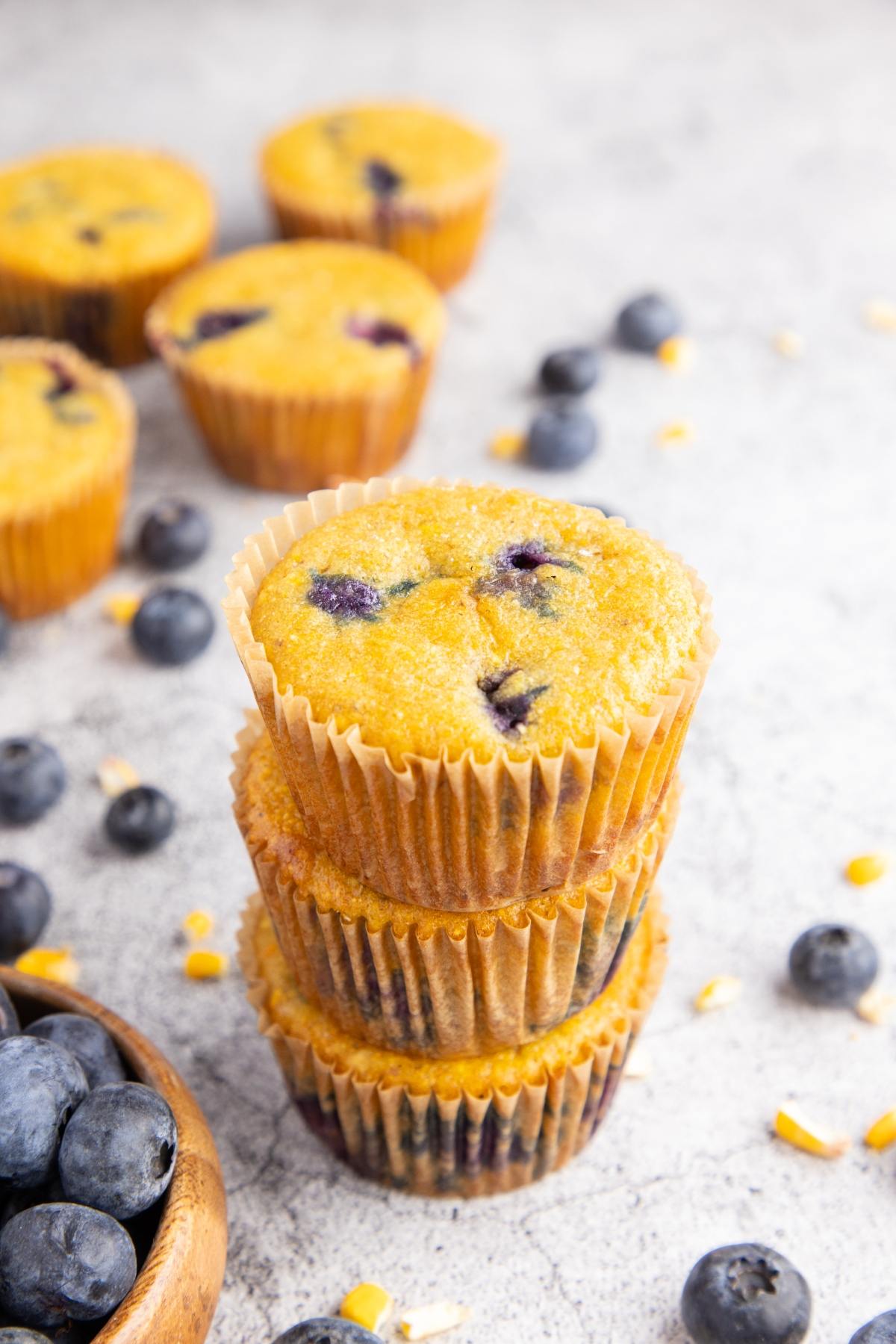 Three blueberry muffins stacked, surrounded by fresh blueberries and scattered muffin pieces on a light surface.