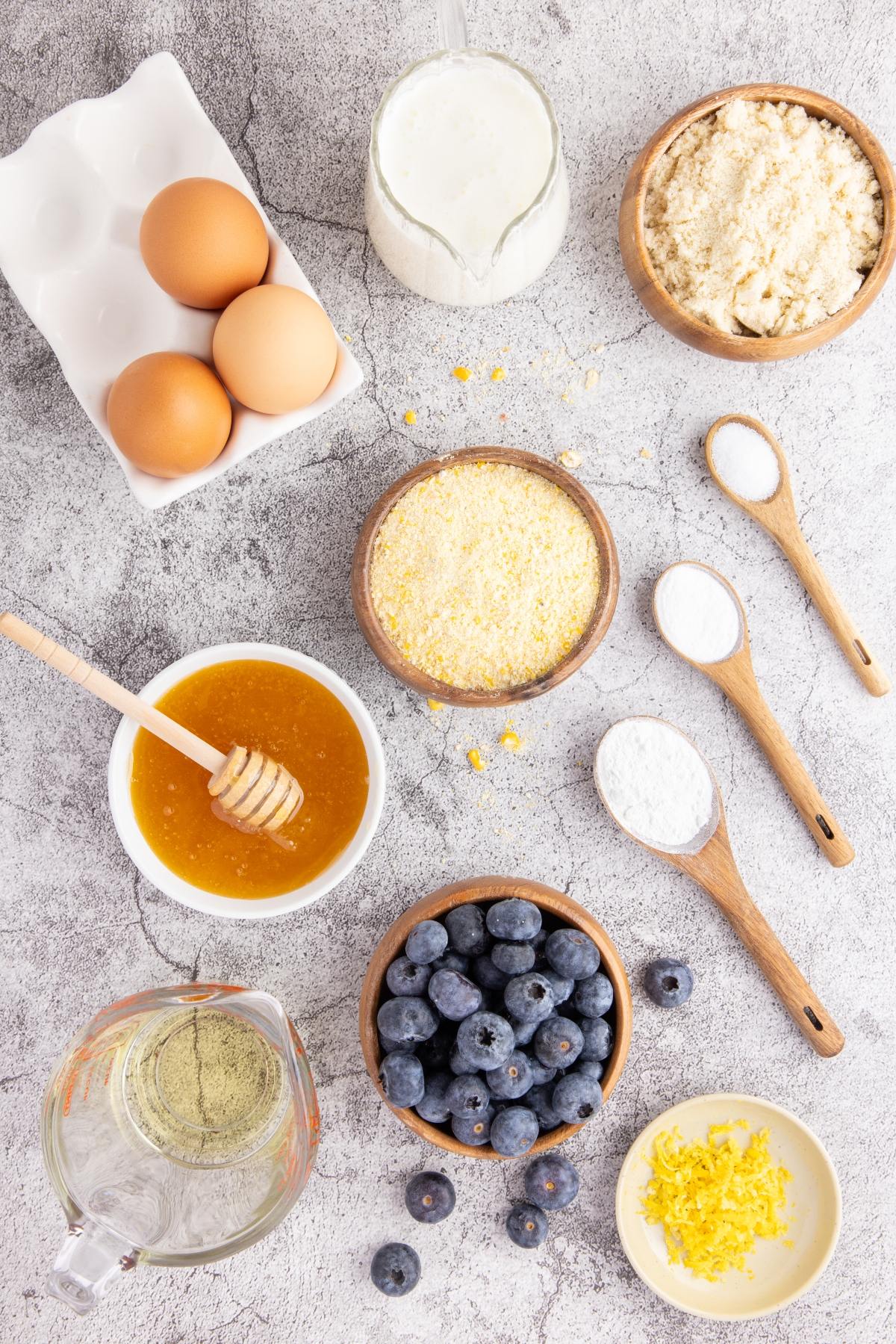 Baking ingredients arranged on a countertop: eggs, milk, honey, blueberries, flour, sugar, and lemon zest.
