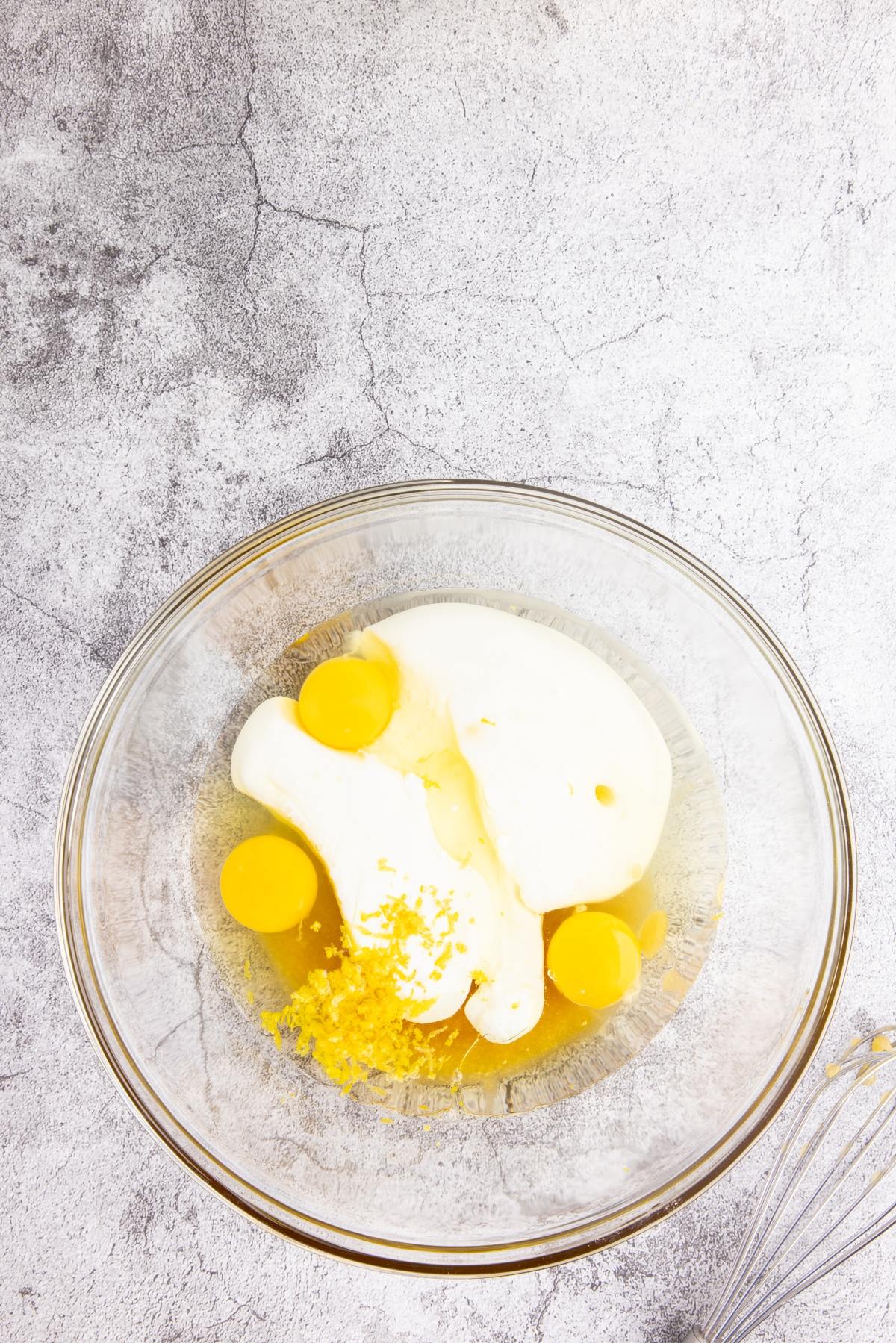 A glass bowl with eggs, sugar, and lemon zest on a gray textured surface next to a metal whisk.