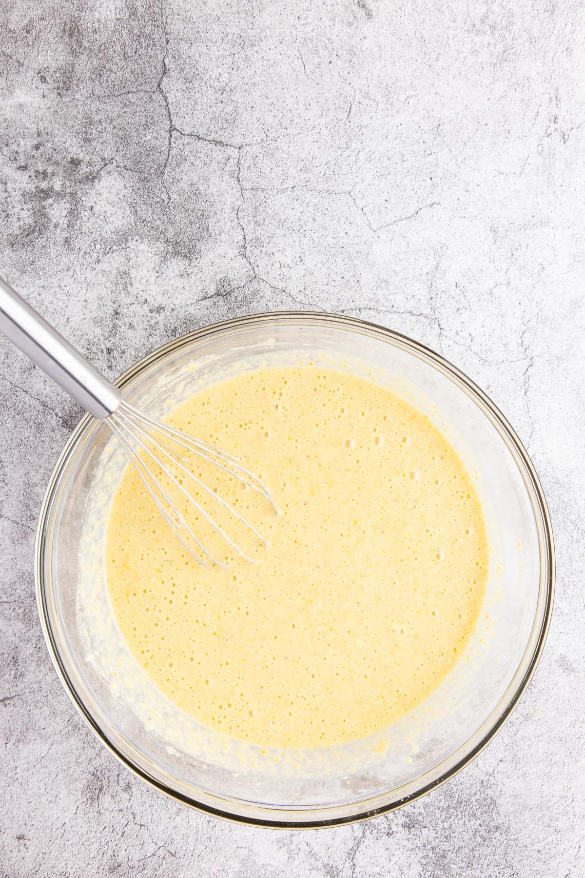A glass bowl of yellow batter with a metal whisk on a light gray textured surface.