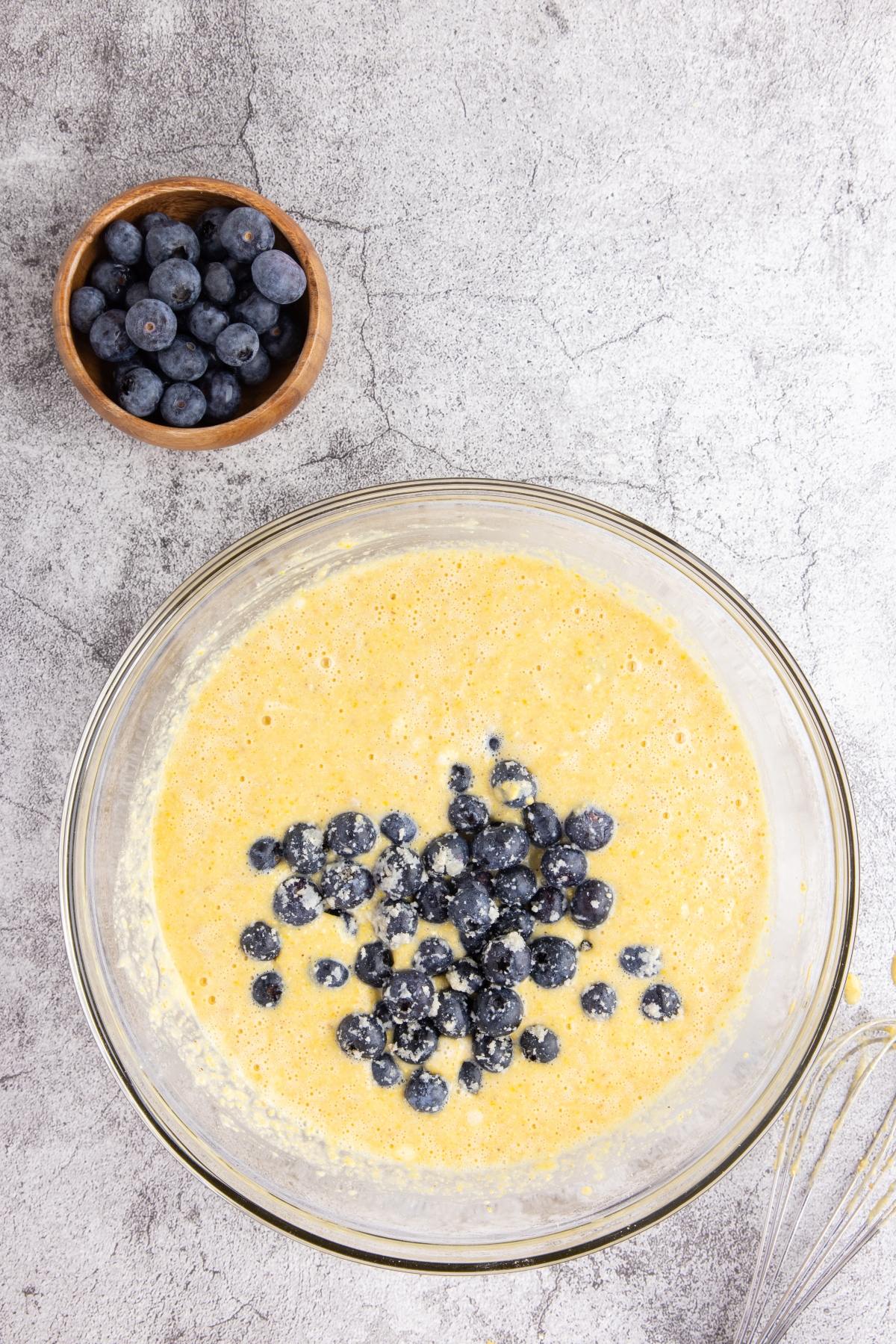 A bowl of batter with blueberries mixed in, and a small bowl of fresh blueberries beside it on a gray surface.
