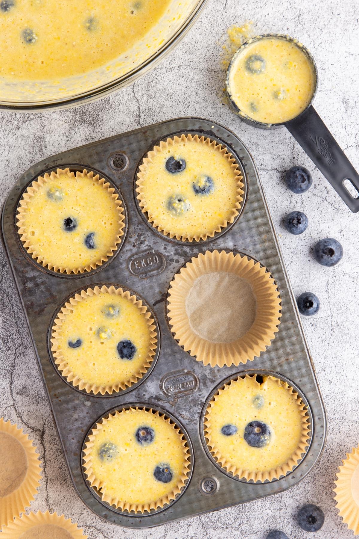 Muffin tin with paper liners filled with blueberry muffin batter, fresh blueberries nearby.