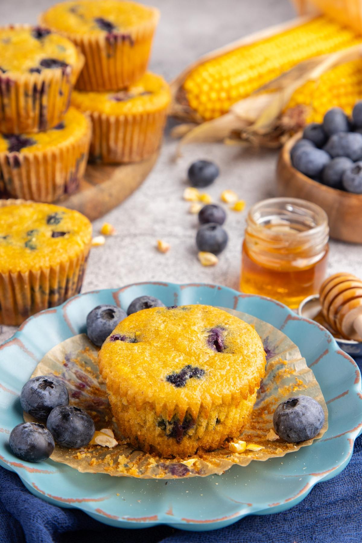 A blueberry corn muffin on a plate with fresh blueberries, honey, and corn in the background.