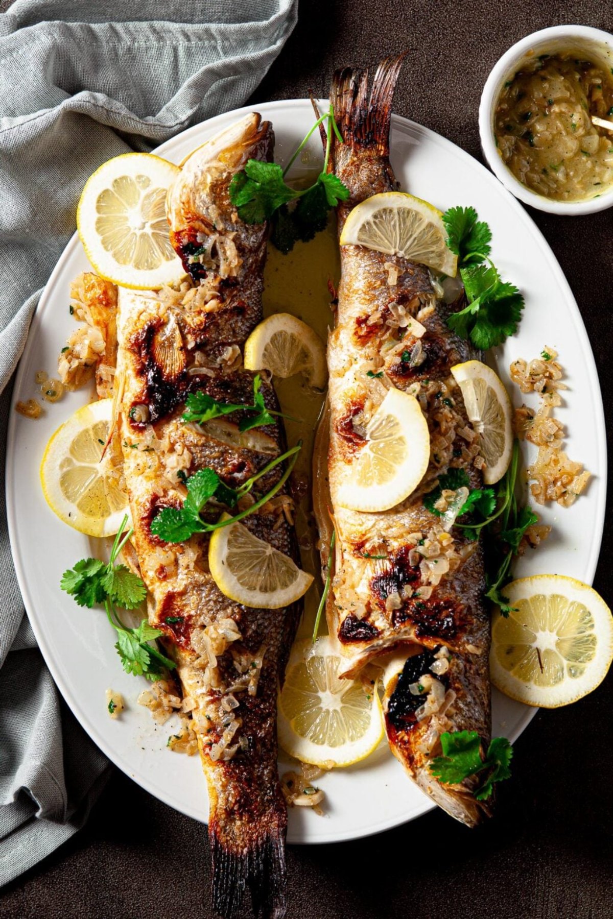 Two whole roasted fish garnished with lemon slices, cilantro, and crispy shallots on a white platter—perfect for adding to your collection of Italian dinner recipes.