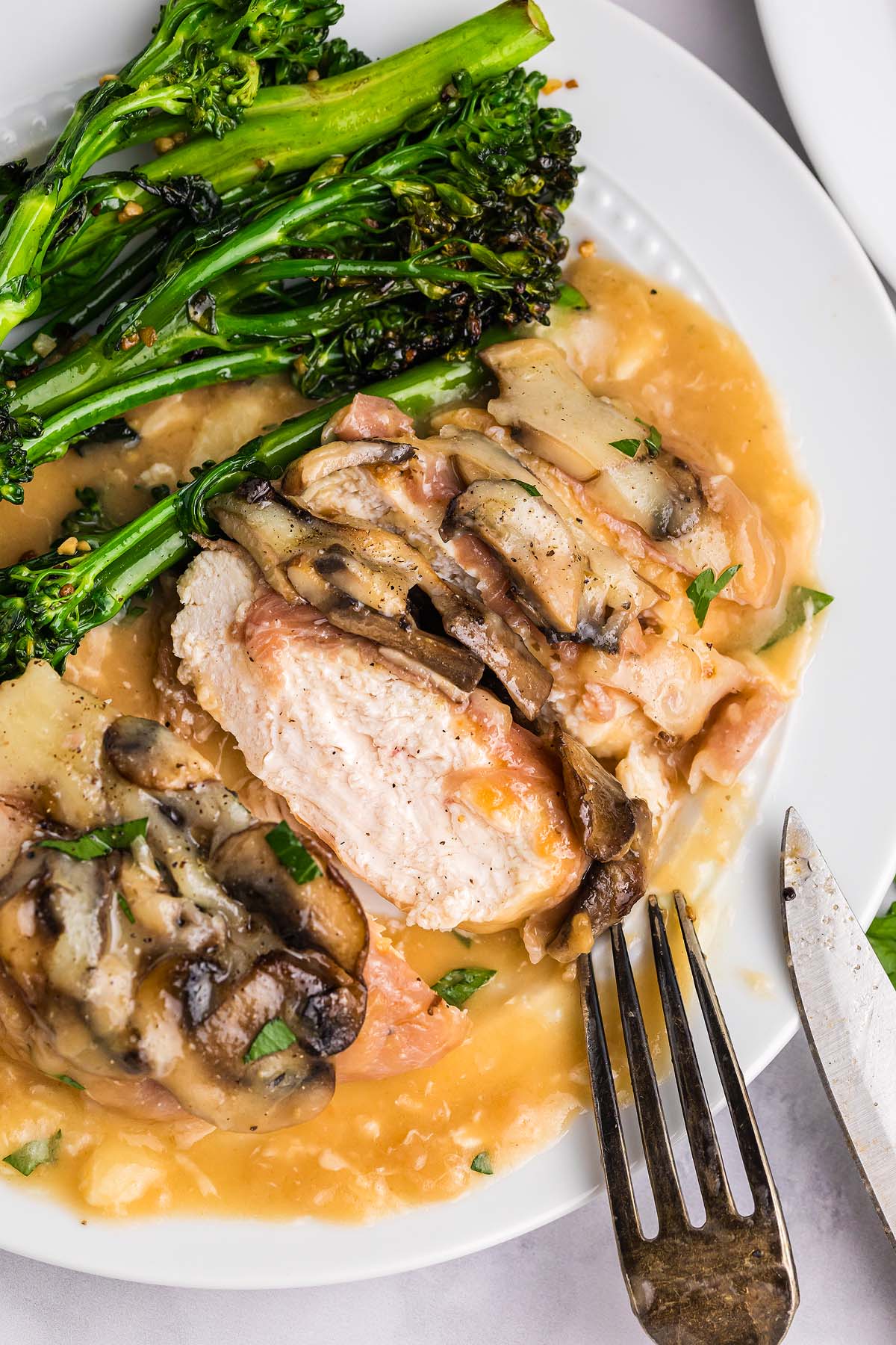 Sliced chicken Valdostana with mushrooms and sauce, served with broccolini on a white plate, next to a fork and knife.