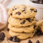 A stack of Chocolate Chip Pudding Cookies rests on parchment paper, with extra chocolate chips and a bowl in the background.