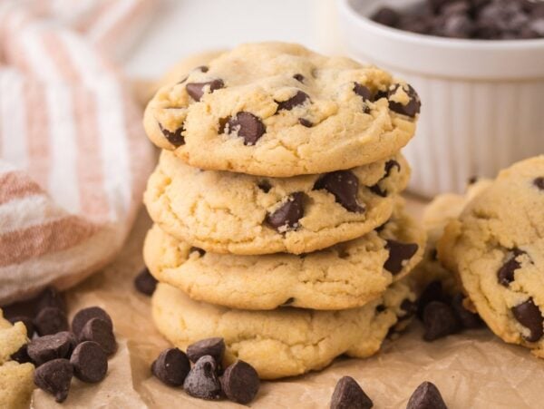 A stack of Chocolate Chip Pudding Cookies rests on parchment paper, with extra chocolate chips and a bowl in the background.