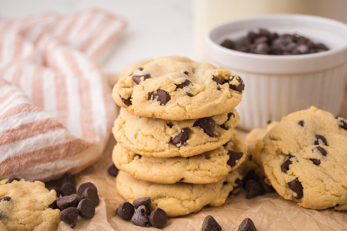 A stack of Chocolate Chip Pudding Cookies on parchment paper, with chocolate chips and a striped towel nearby.