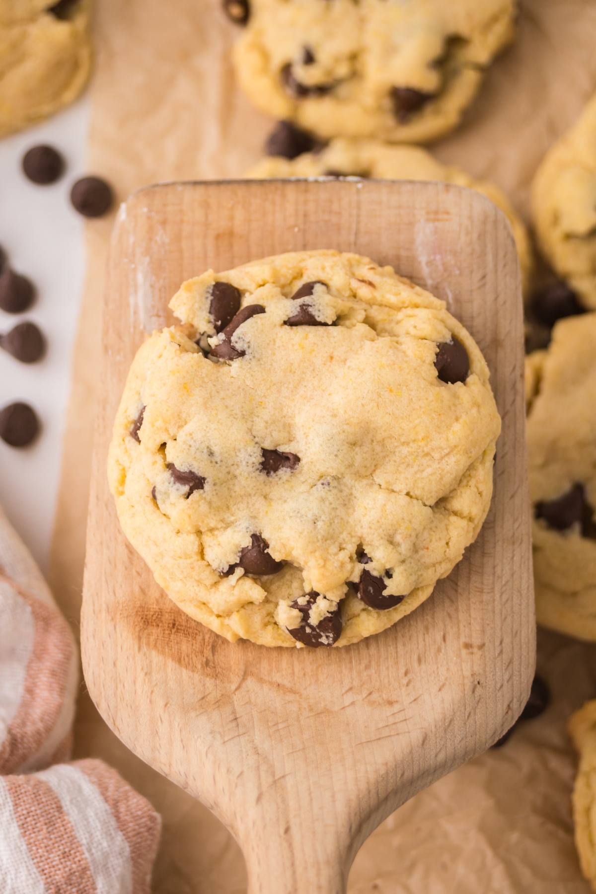 A thick chocolate chip cookie on a wooden spatula, surrounded by more cookies and chocolate chips.
