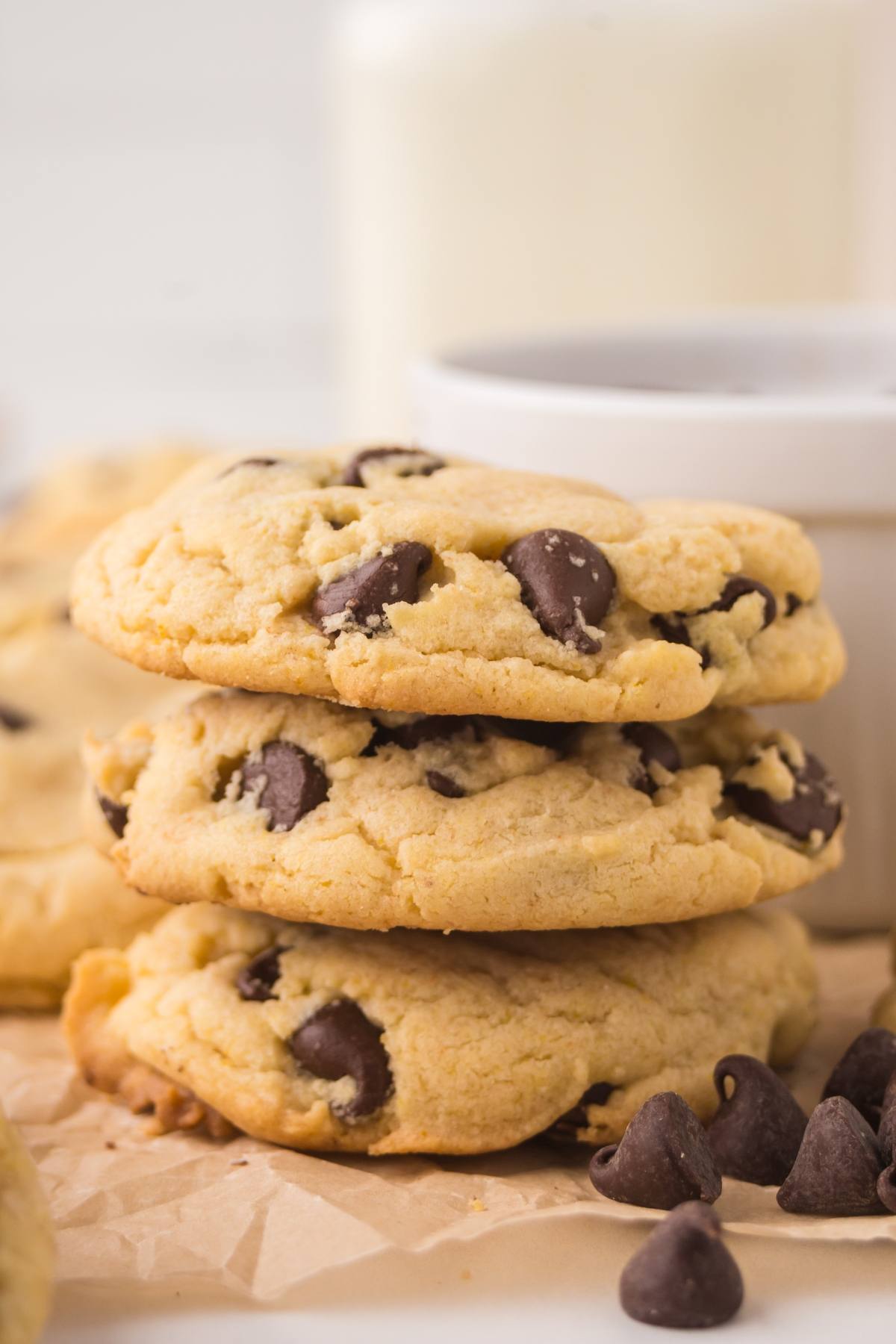 Three chocolate chip cookies stacked on parchment paper with chocolate chips and a cup in the background.