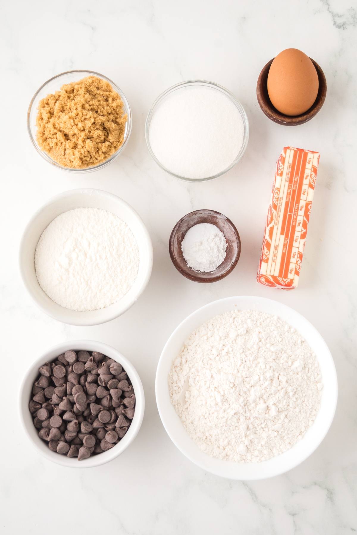 Baking ingredients on a white surface: sugars, flour, baking soda, butter, egg, and chocolate chips.