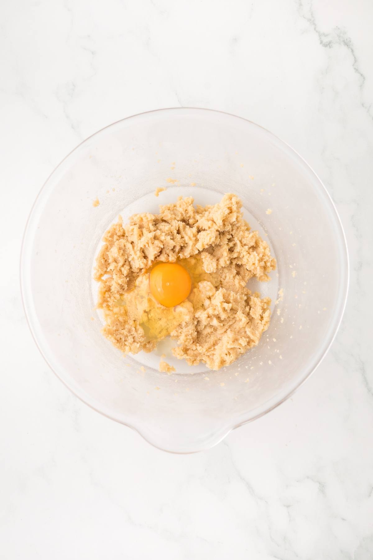 A clear bowl with cookie dough and a raw egg on a white marble surface.