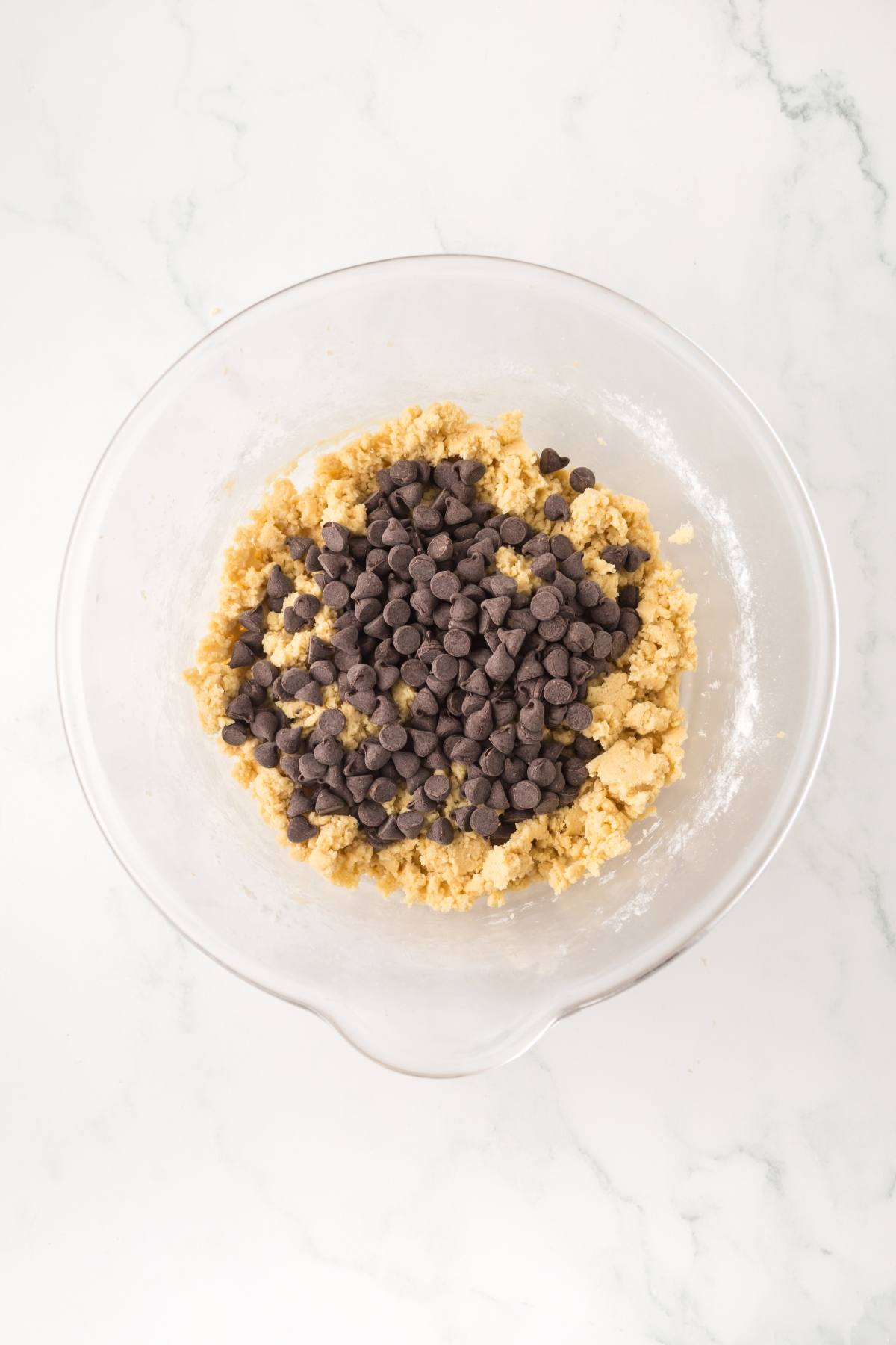 A clear bowl of cookie dough topped with chocolate chips on a white marble surface.