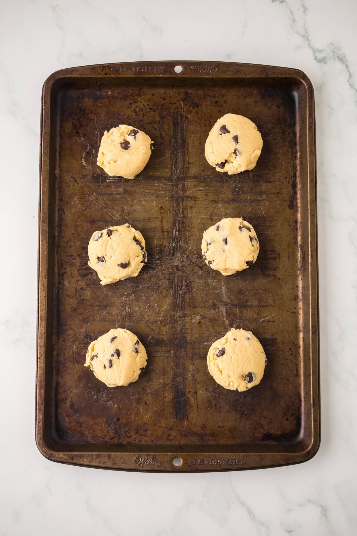 Six unbaked chocolate chip cookie dough balls on a worn metal baking sheet.