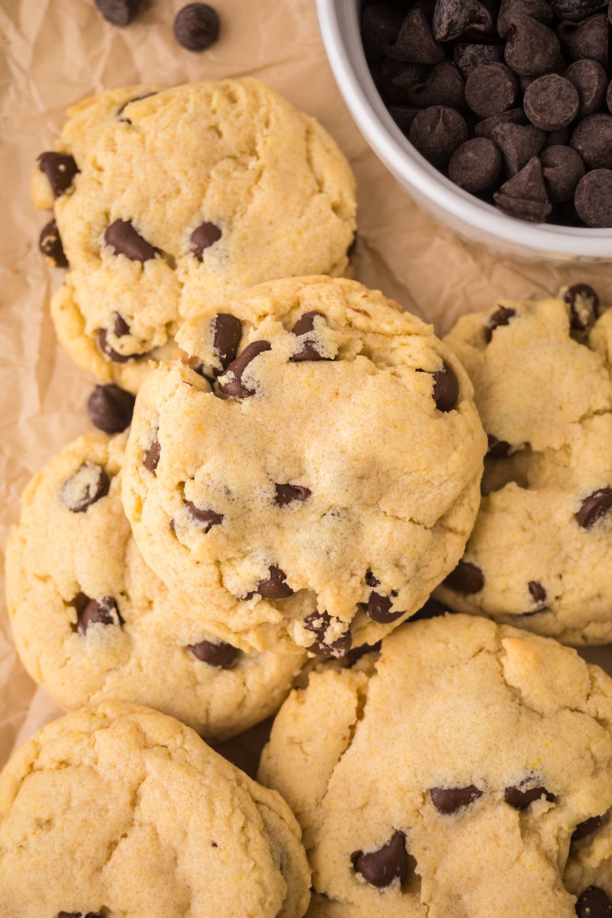 Chocolate chip cookies piled on brown parchment paper next to a white bowl filled with chocolate chips.