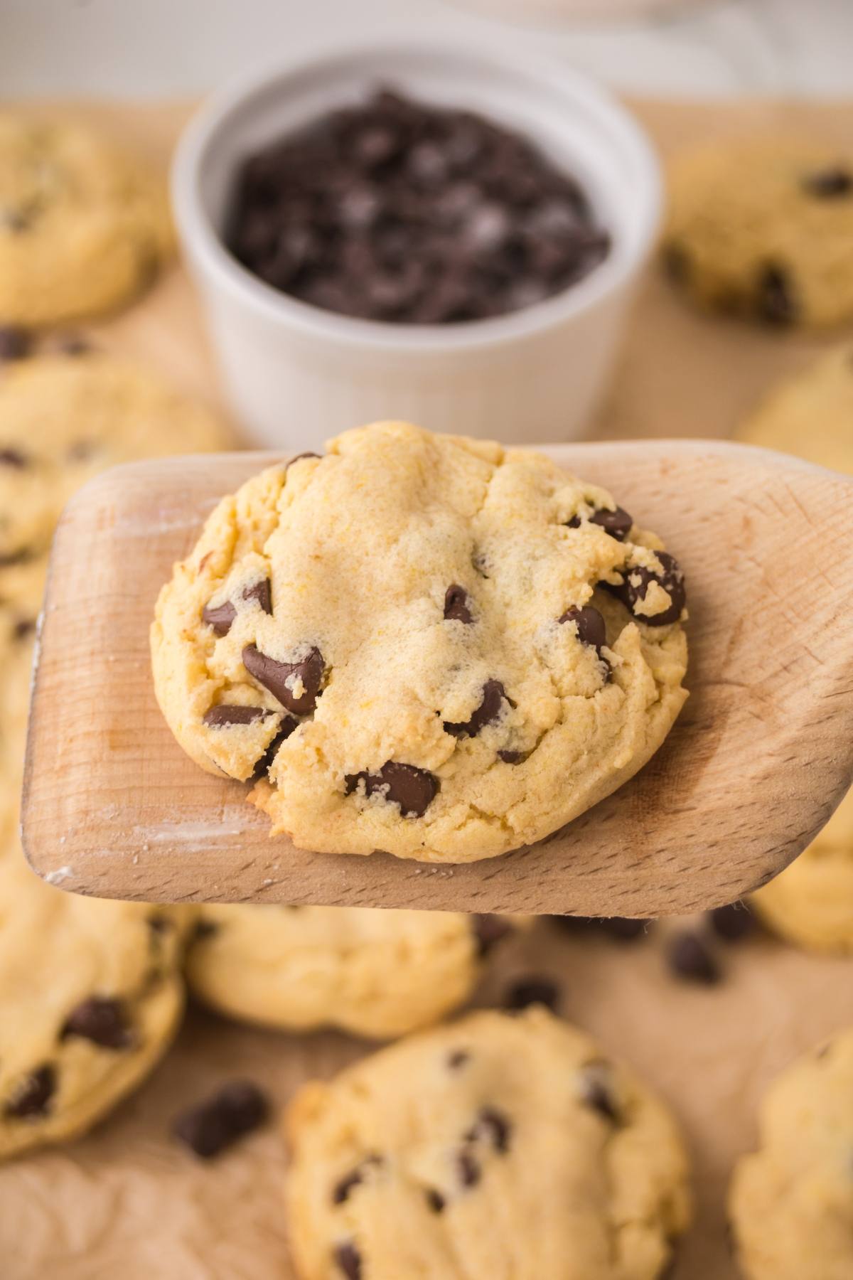 A wooden spatula holding a chocolate chip cookie above several cookies and a bowl of chocolate chips.