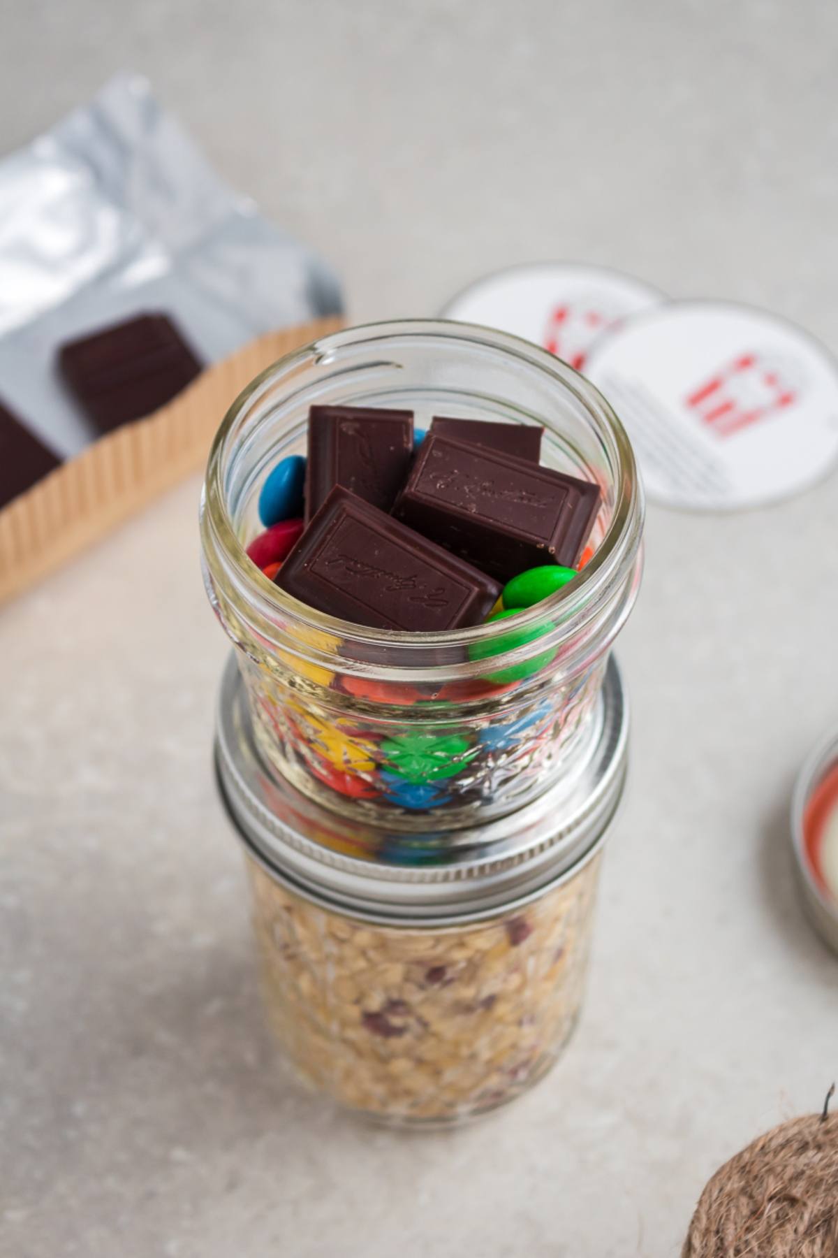 Mason jar filled with chocolate pieces and colorful candies, sitting on top of another jar with oats—perfect for creating your own Chocolate M&M Popcorn DIY Gift Kit.