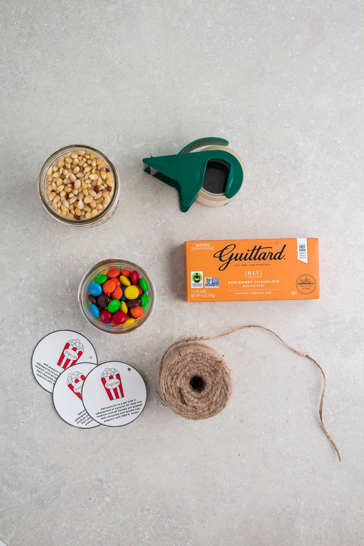 Chocolate M&M Popcorn DIY Gift Kit includes popcorn kernels, M&Ms, a chocolate bar, tape, twine, and two popcorn-themed round gift tags on a gray surface.
