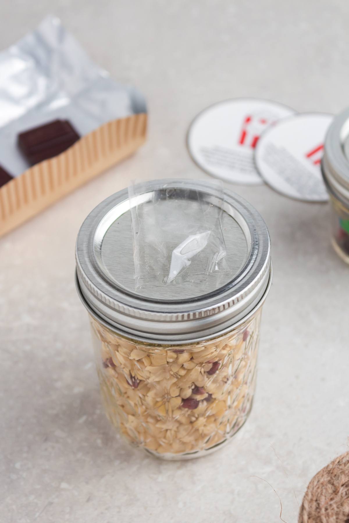A mason jar filled with oats and seeds, topped with a sealed lid and a plastic-wrapped scoop, makes a charming Chocolate M&M Popcorn DIY Gift Kit.