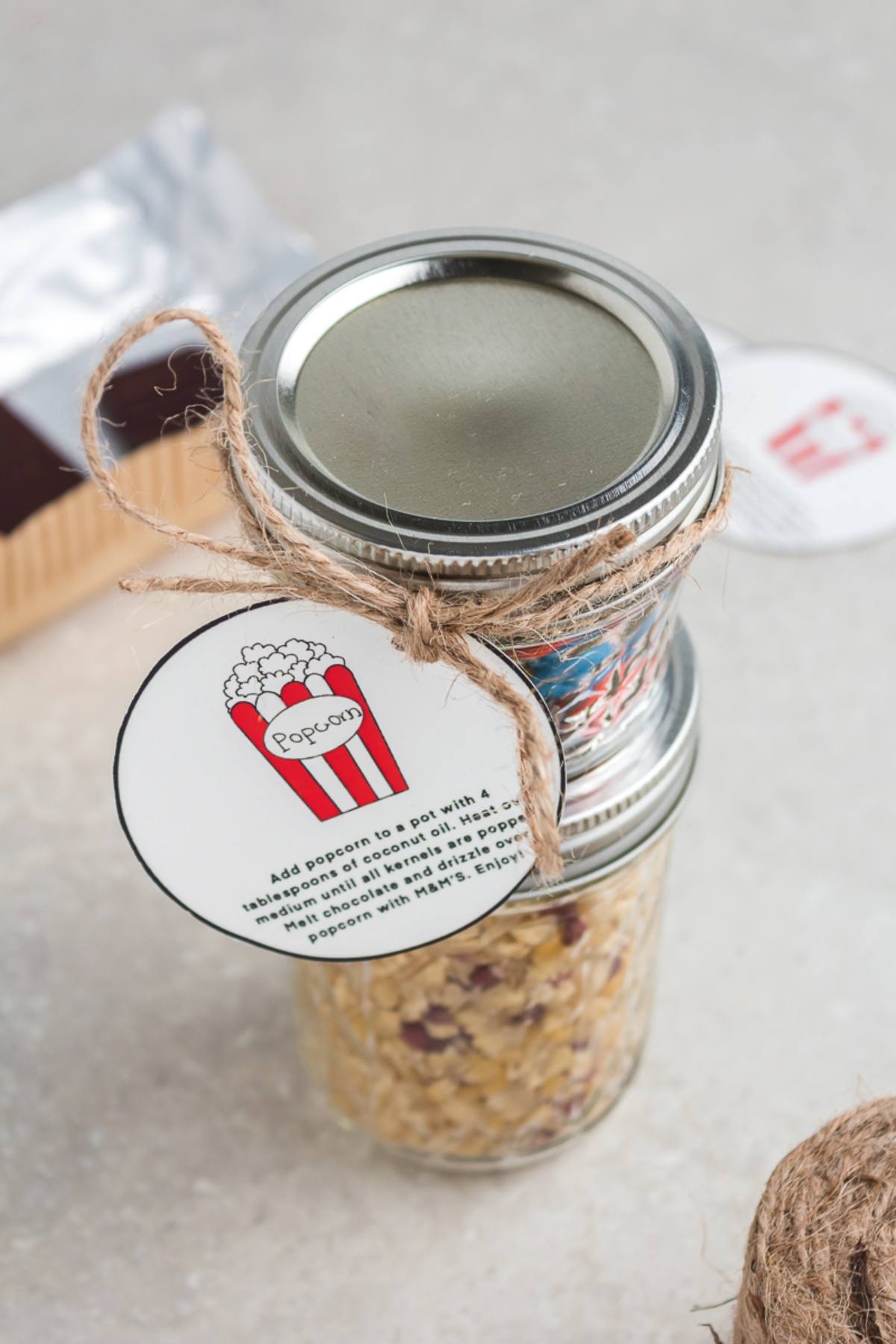 A glass jar filled with popcorn kernels, tied with twine and a round Chocolate M&M Popcorn DIY Gift Kit label, on a light surface.