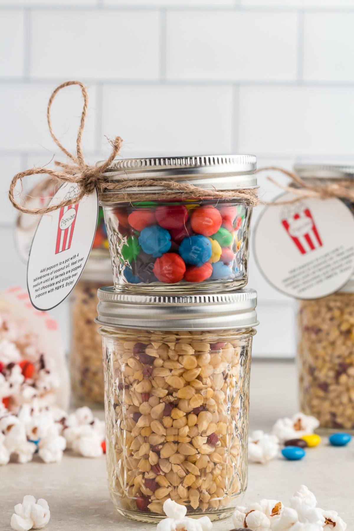 Two mason jars stacked, one with popcorn kernels and one with colorful Chocolate M&M candies, tied with twine and a gift tag— the perfect Popcorn DIY Gift Kit.