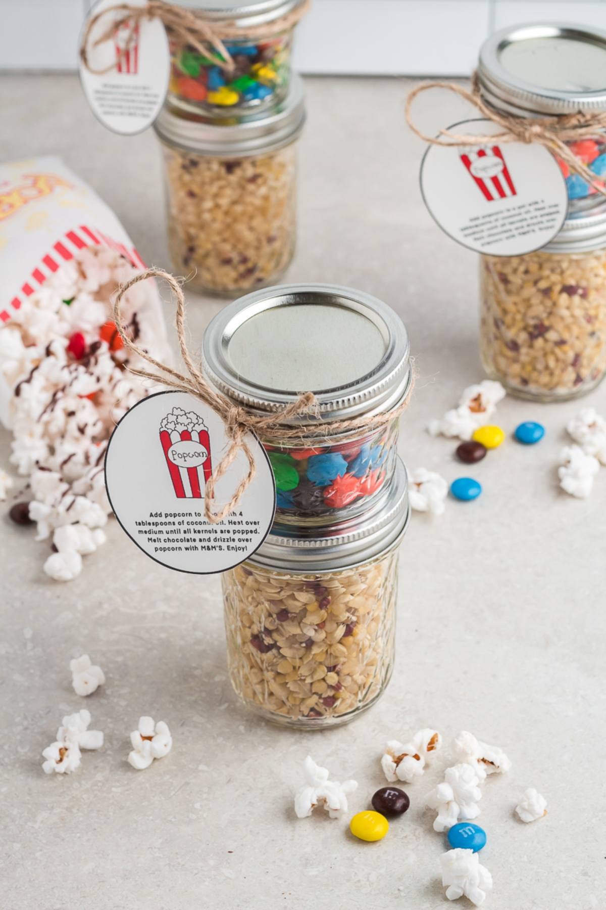 Three jars stacked with popcorn kernels and candy, tied with tags, surrounded by popcorn and colorful candies—a delightful Chocolate M&M Popcorn DIY Gift Kit.