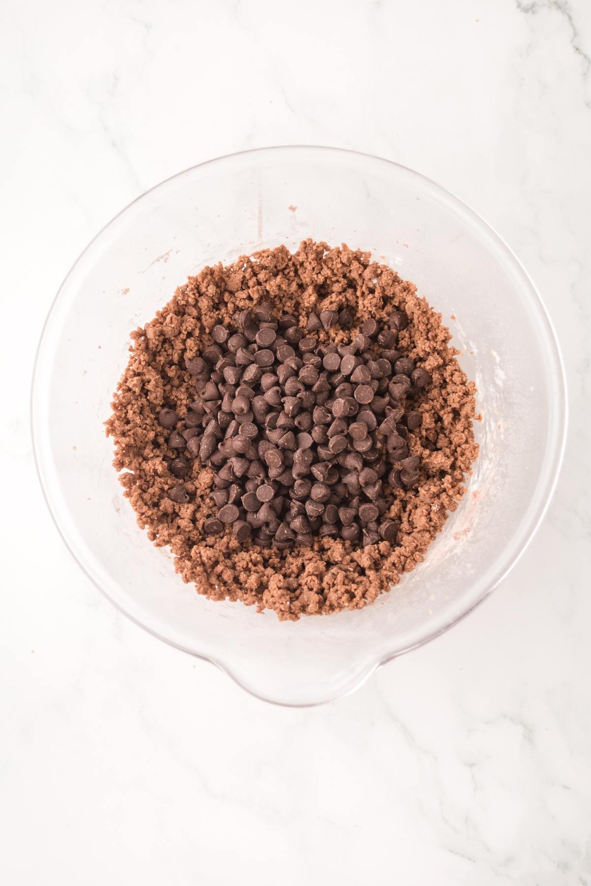 A glass bowl with chocolate cookie dough and chocolate chips on a white marble surface.