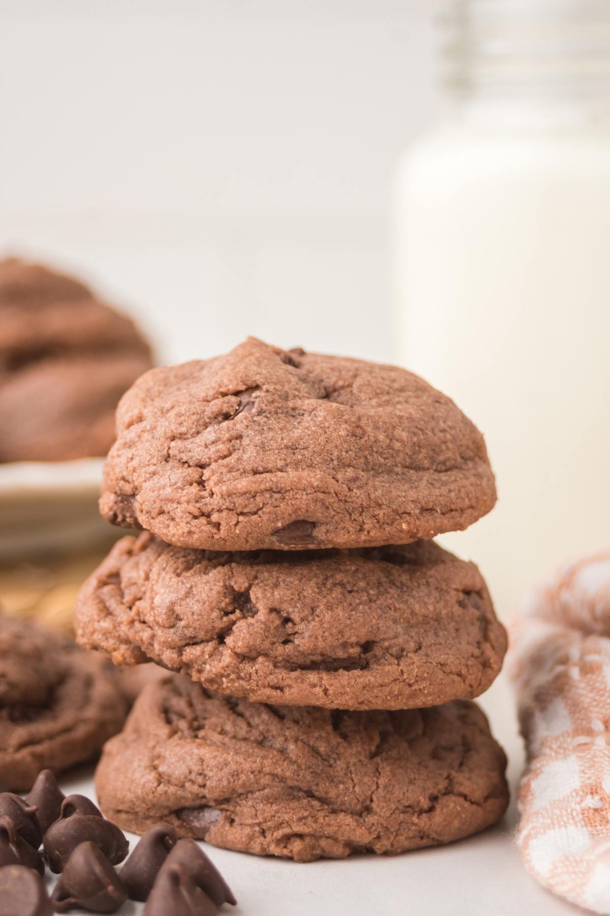 Three chocolate cookies stacked with chocolate chips nearby and a bottle of milk in the background.
