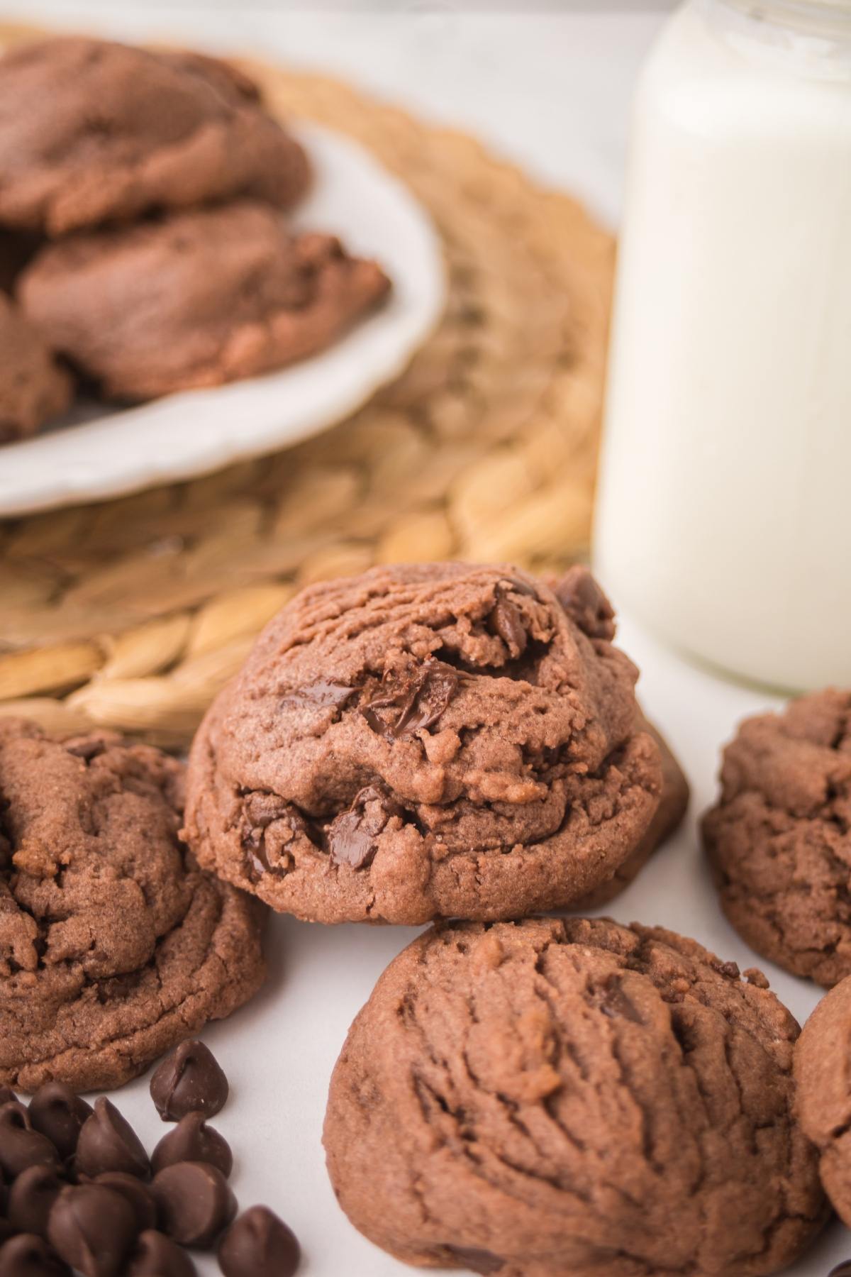 Chocolate cookies with chocolate chips next to a glass of milk and more cookies on a plate in the background.